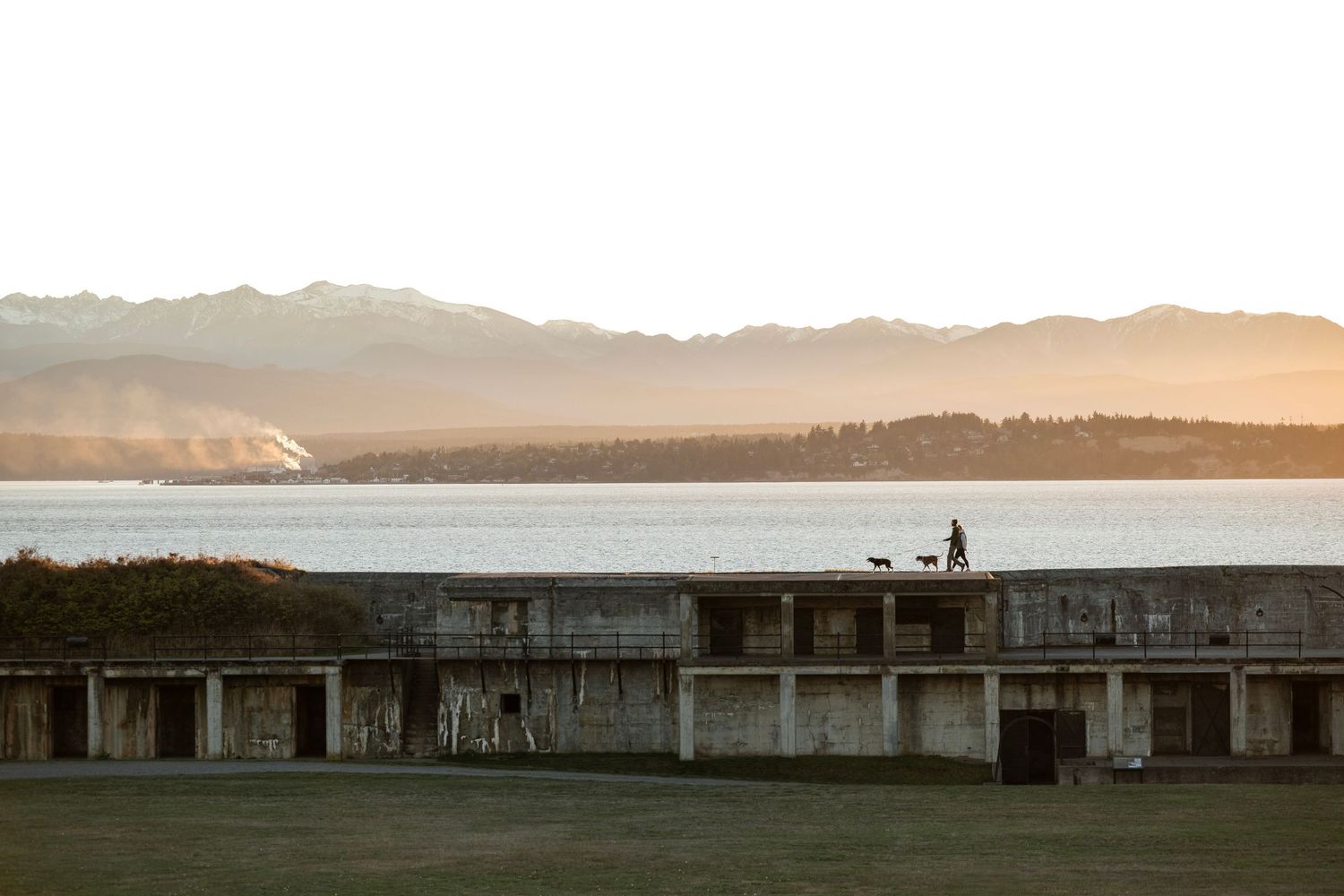 Person walking dogs on historic fort ruins at sunset with mountain and water views in background.