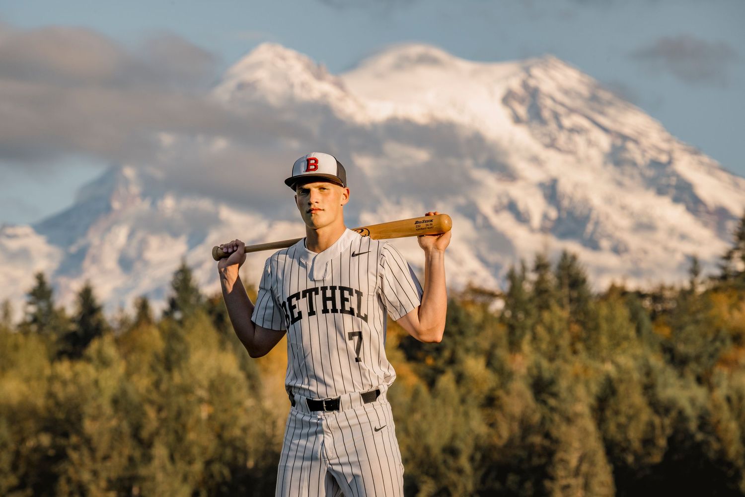 Bethel baseball player #7 poses with bat on shoulder, snow-capped Mount Rainier in background.
