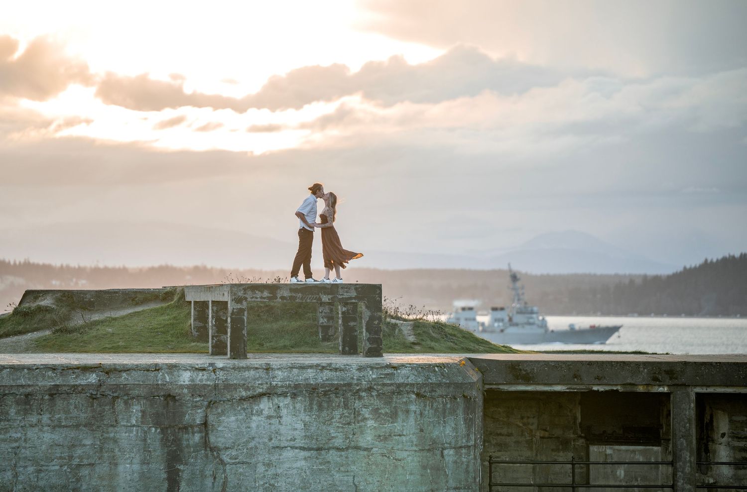Couple sharing a romantic kiss on a concrete fort overlooking a bay with a ship at sunset.