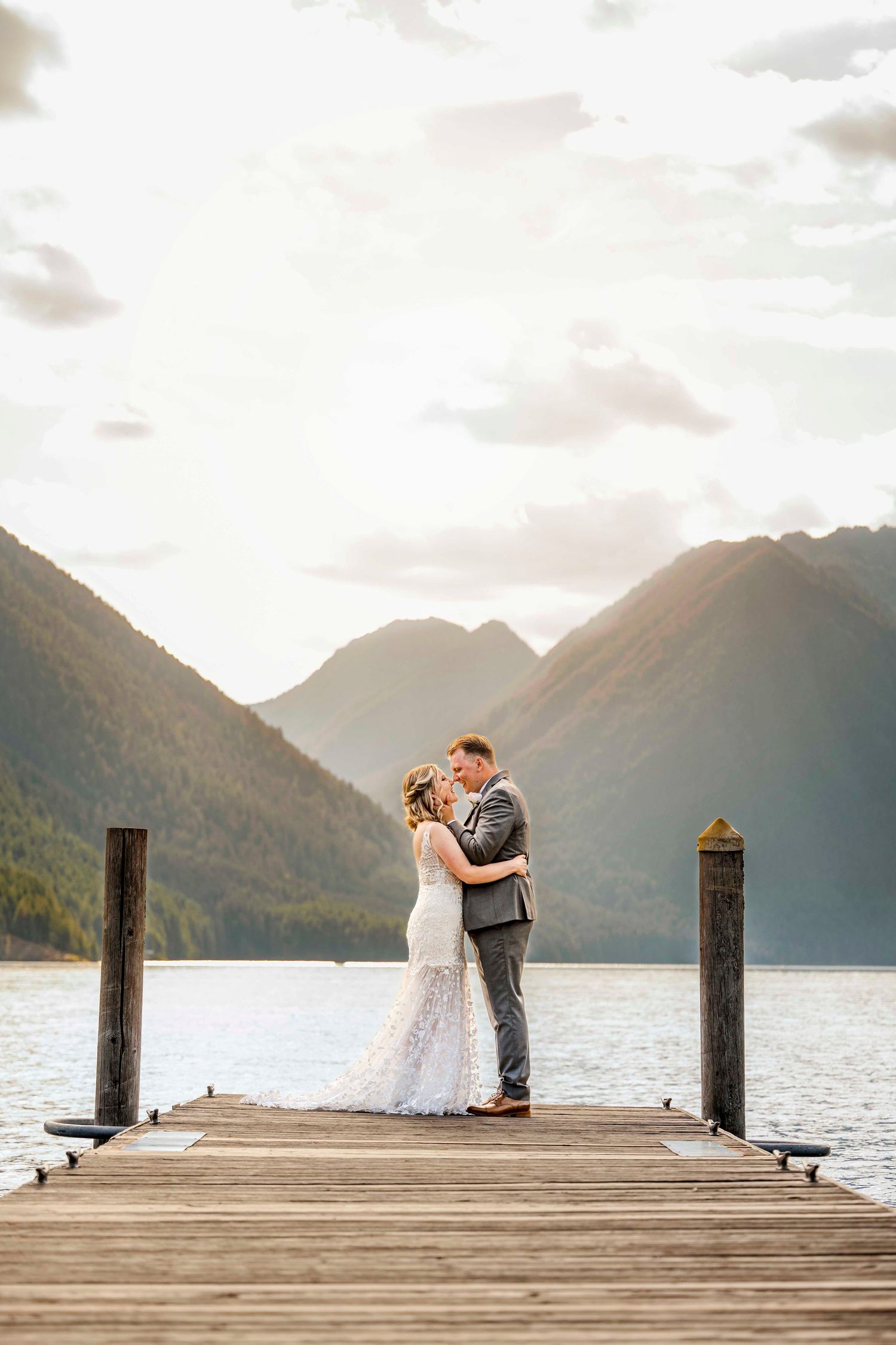 Bride and groom sharing romantic kiss on wooden dock with mountain lake backdrop at sunset.