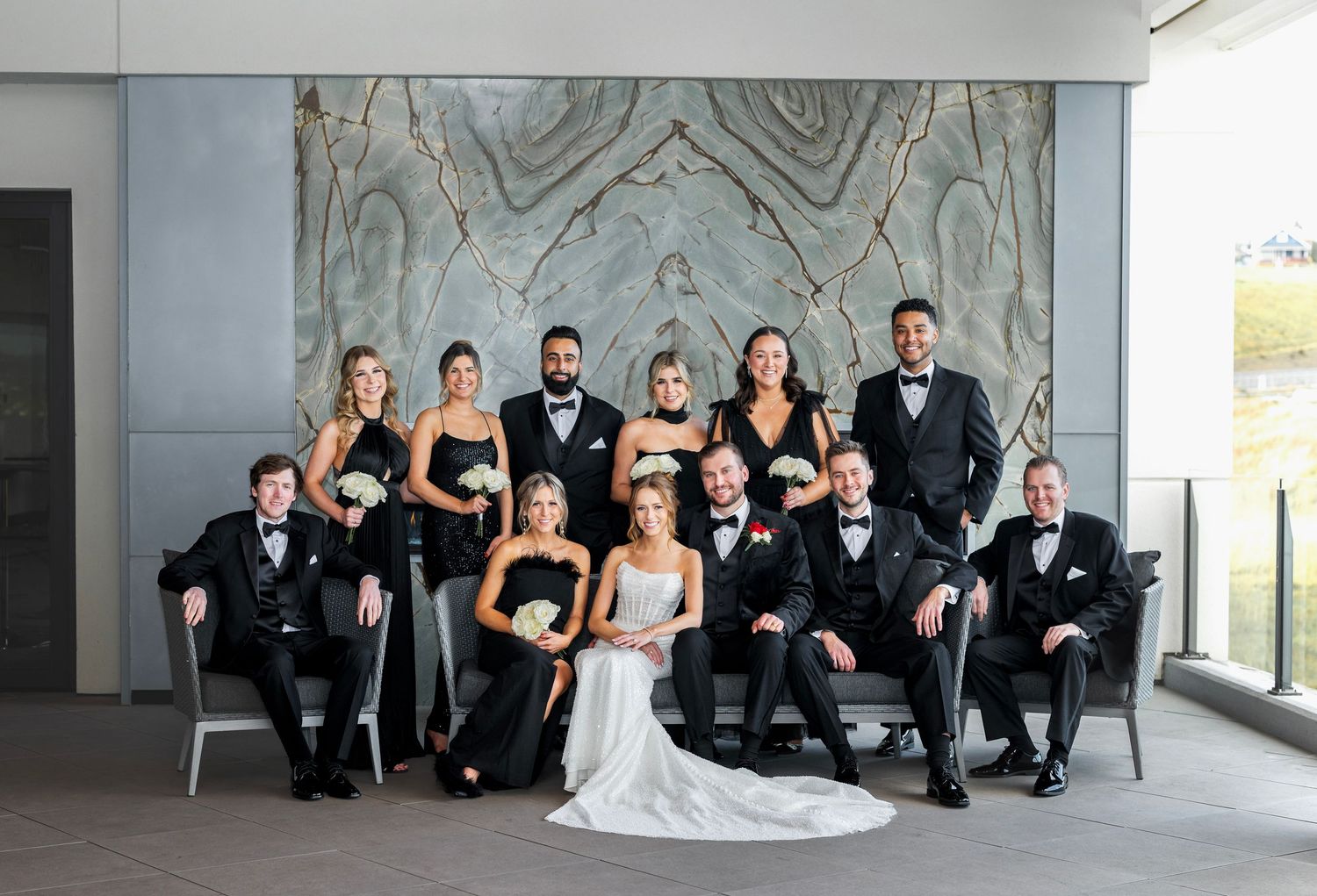 Wedding party posing elegantly in black formal attire before a marble wall backdrop.