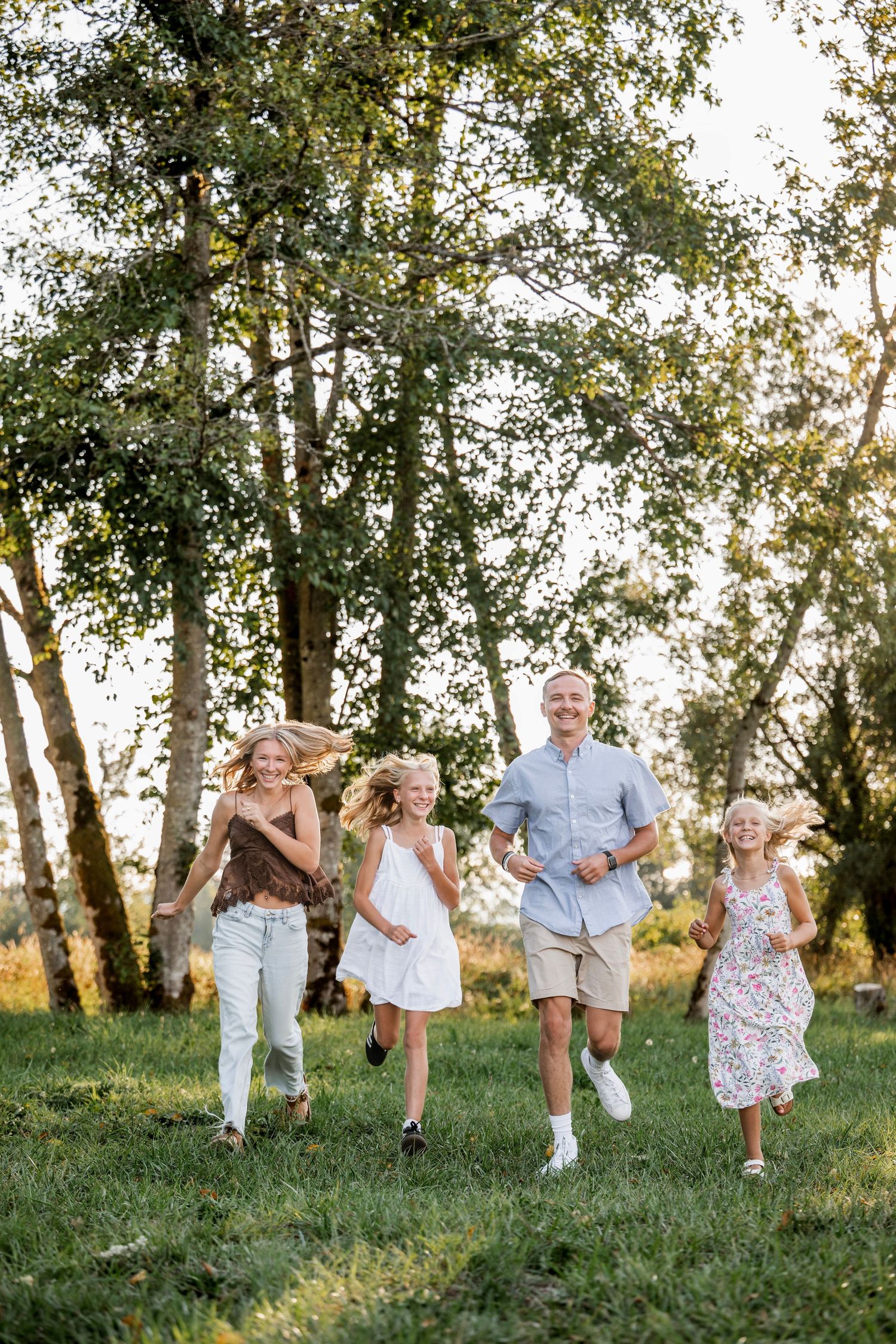 A happy family with two adults and three children running and laughing together on a green lawn surrounded by trees.