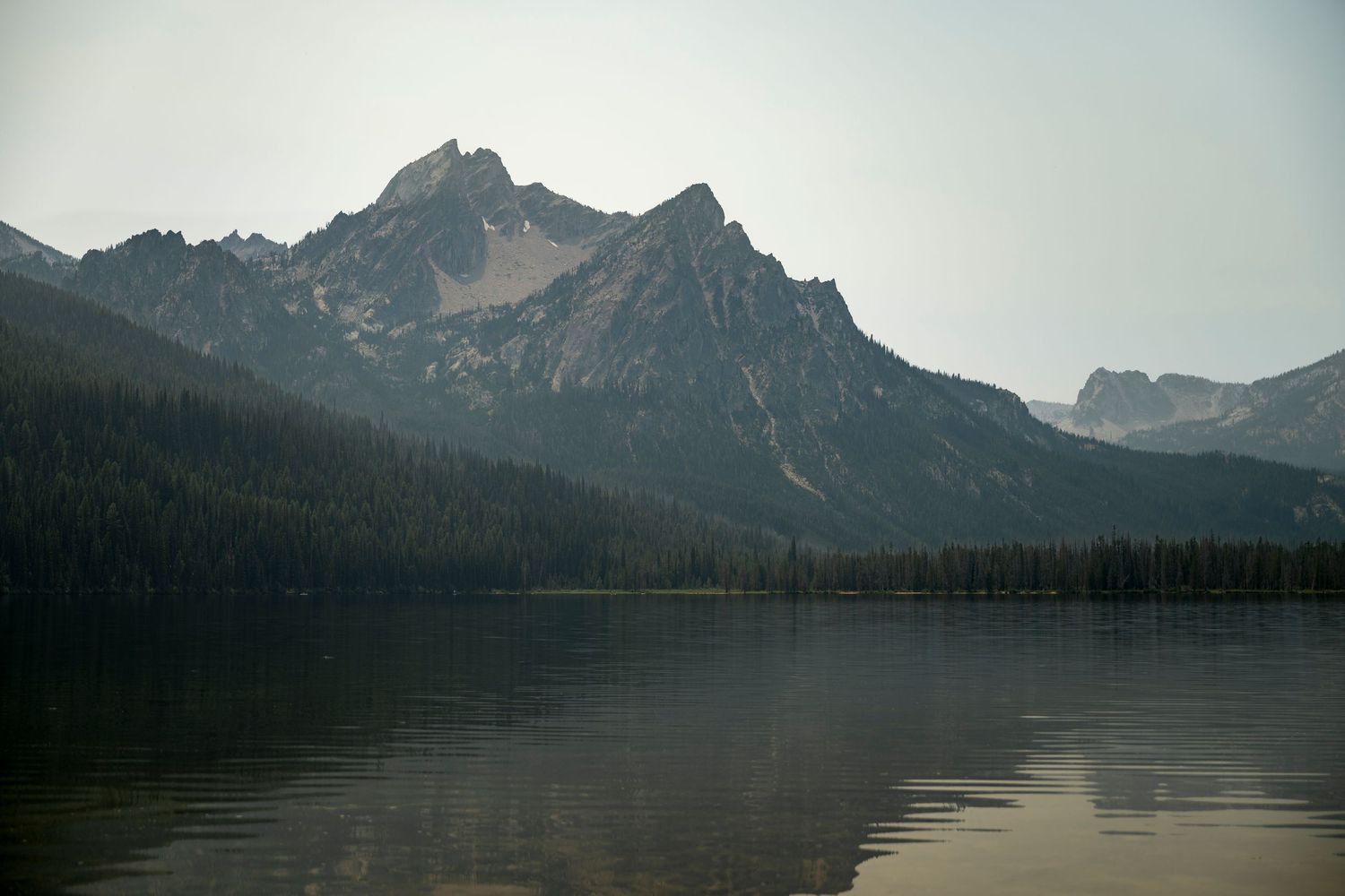 Misty mountain peaks reflected in a calm alpine lake surrounded by dense pine forests.