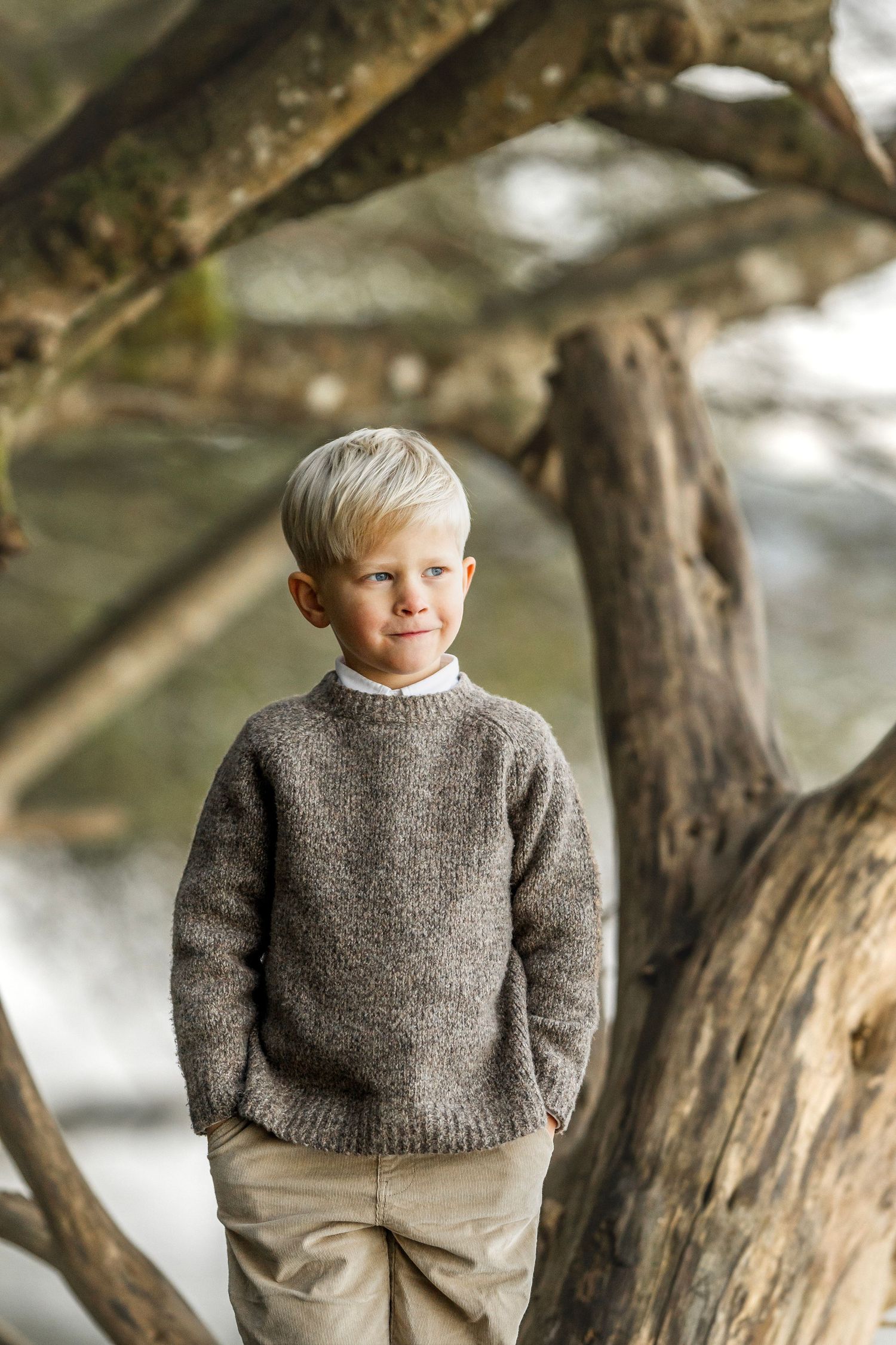 Blonde boy in brown knit sweater stands among tree roots outdoors.