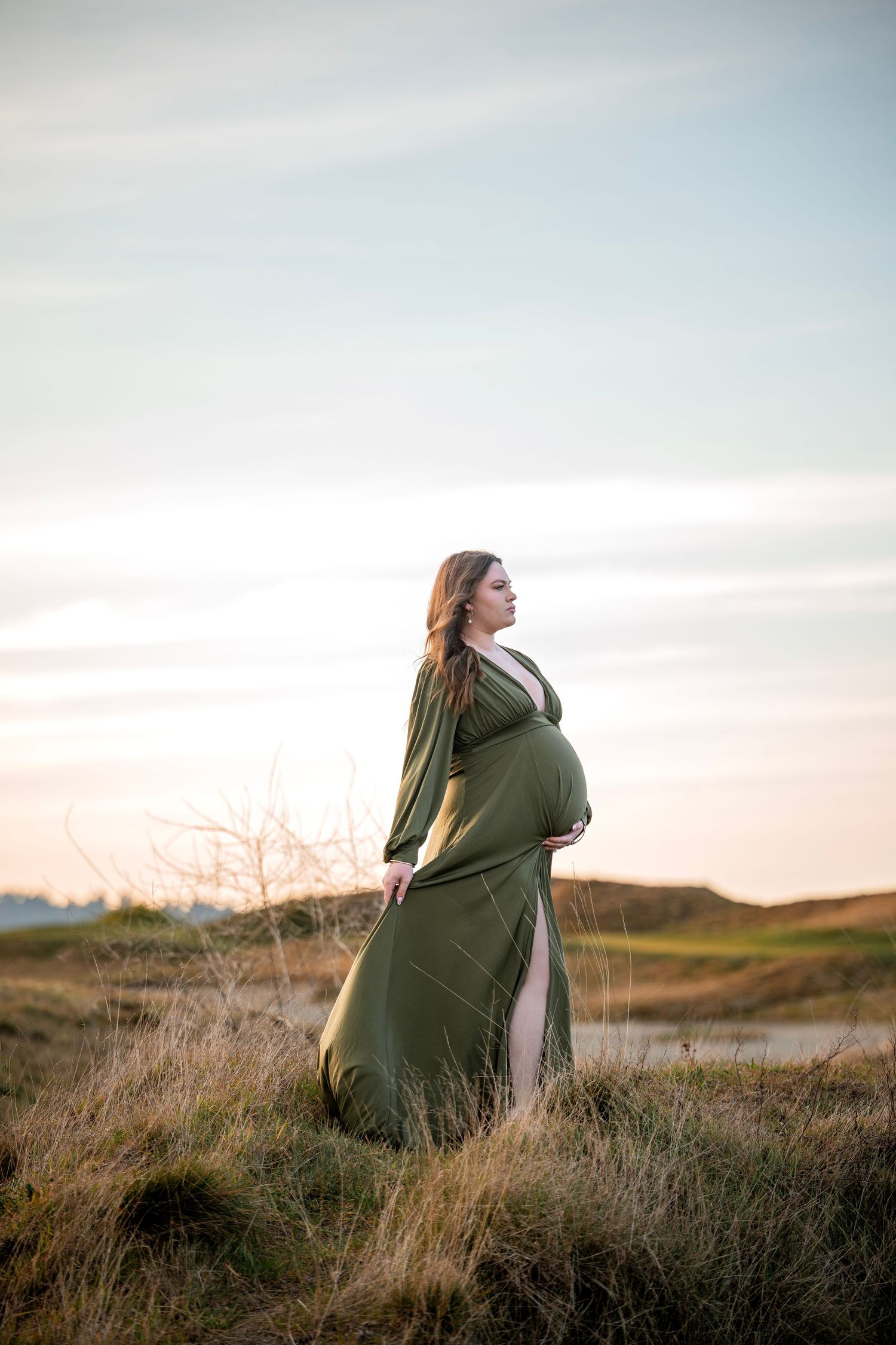 Pregnant woman in an olive green flowing gown standing in a field at sunset.
