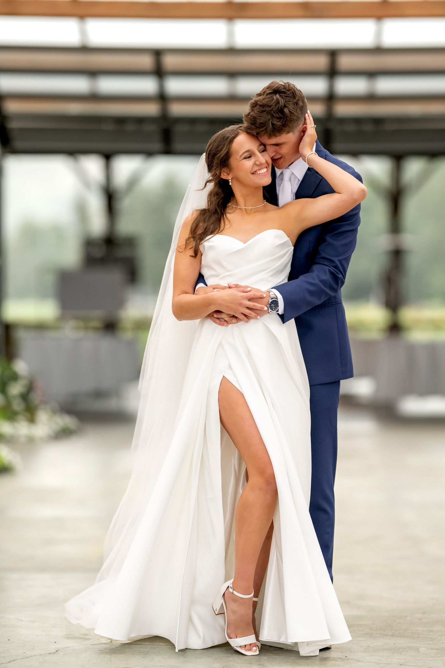 Bride in white gown with slit embraces groom in navy suit during romantic wedding portrait indoors.