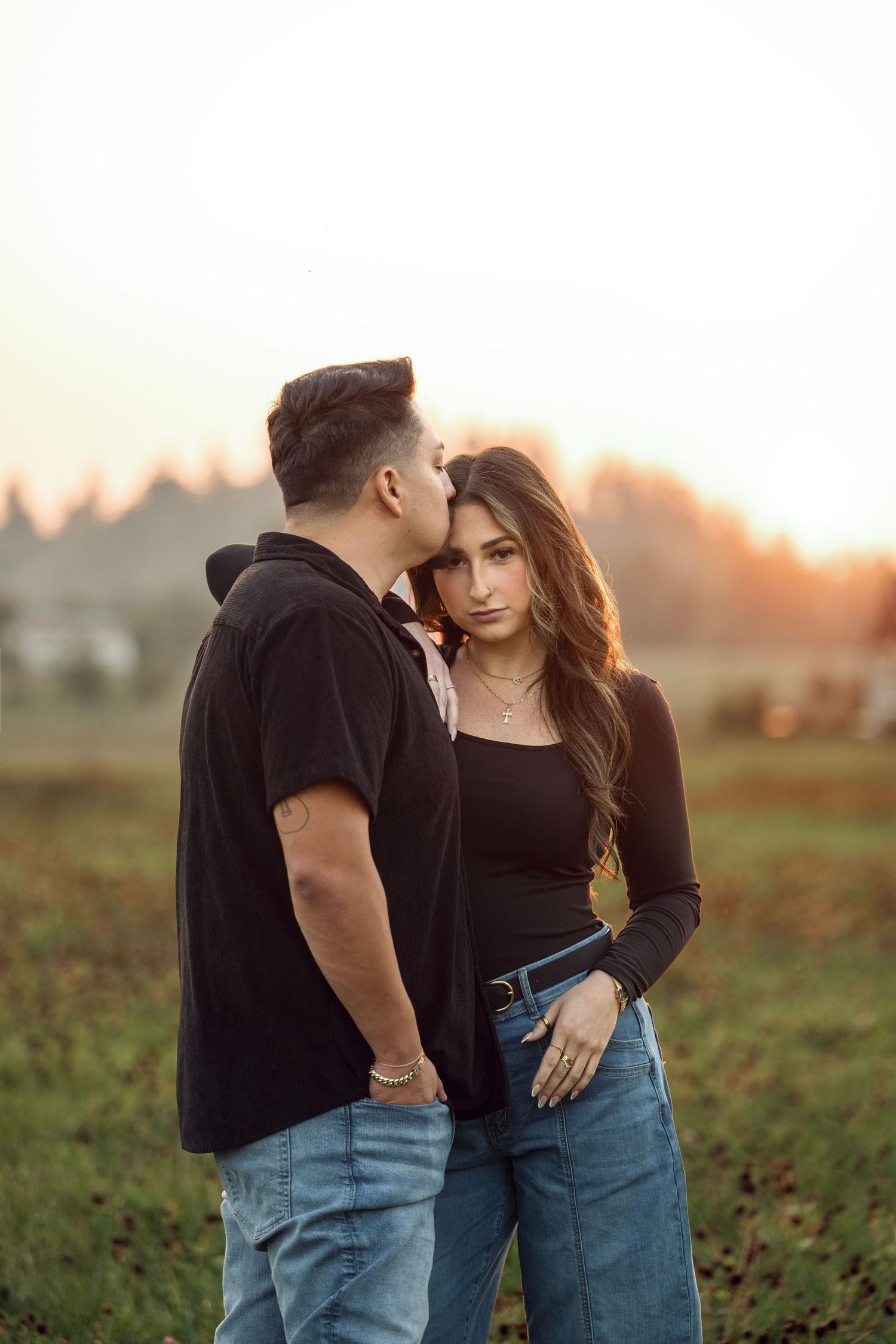 Couple standing in a golden sunset field, man kissing woman's cheek as she gazes forward.
