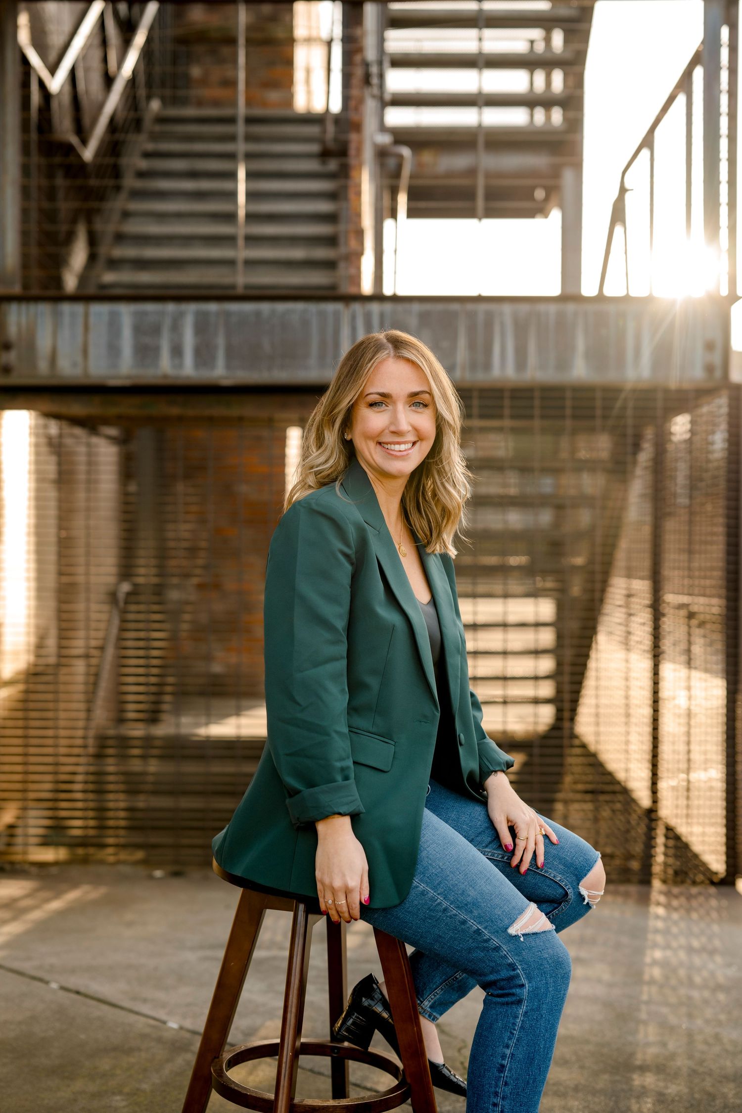 Smiling blonde woman in green blazer and jeans sits on stool with sunlit industrial staircase backdrop.
