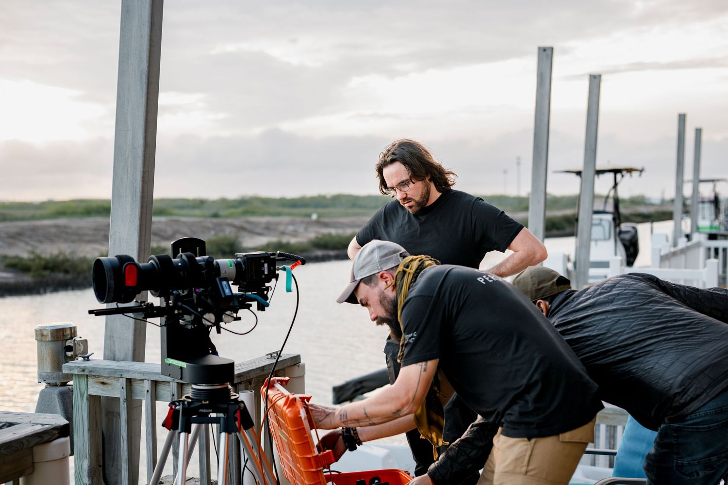Film crew with camera equipment working on a dock near water at dusk during an outdoor production shoot.