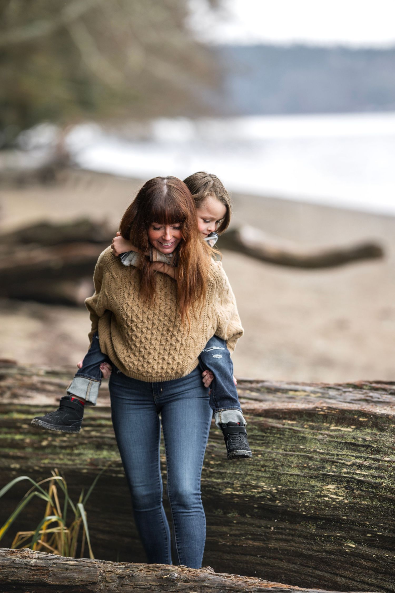 Mother giving young daughter a piggyback ride on a beach log, both smiling in cozy knit sweaters.