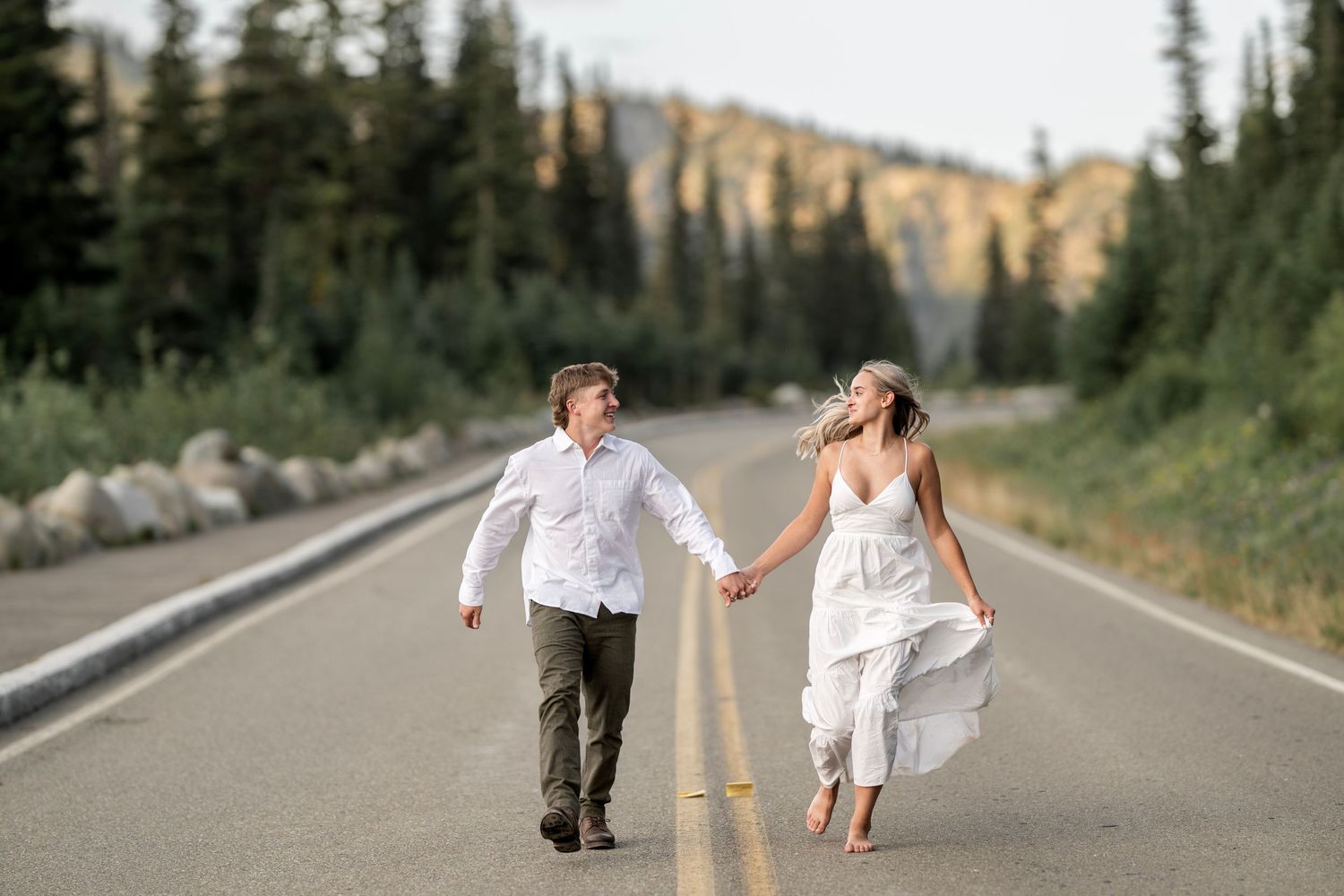 Couple holding hands walking on a mountain road surrounded by pine trees at sunset.