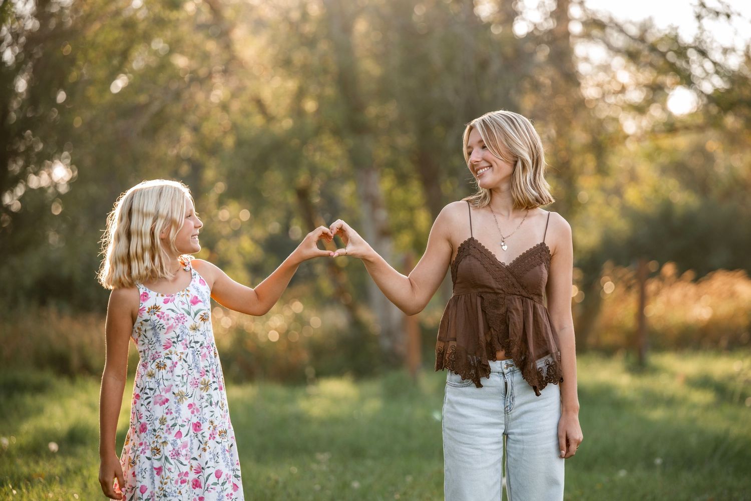 A woman and young girl make a heart shape with their hands outdoors in a sunny green park setting.