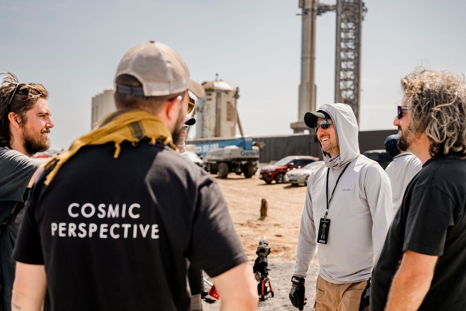 Group of four people talking outdoors near SpaceX rocket launch facility, one wearing Cosmic Perspective shirt.