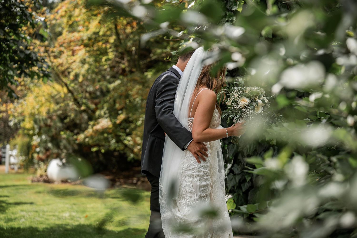 Bride in lace gown with veil walking with groom through lush garden.