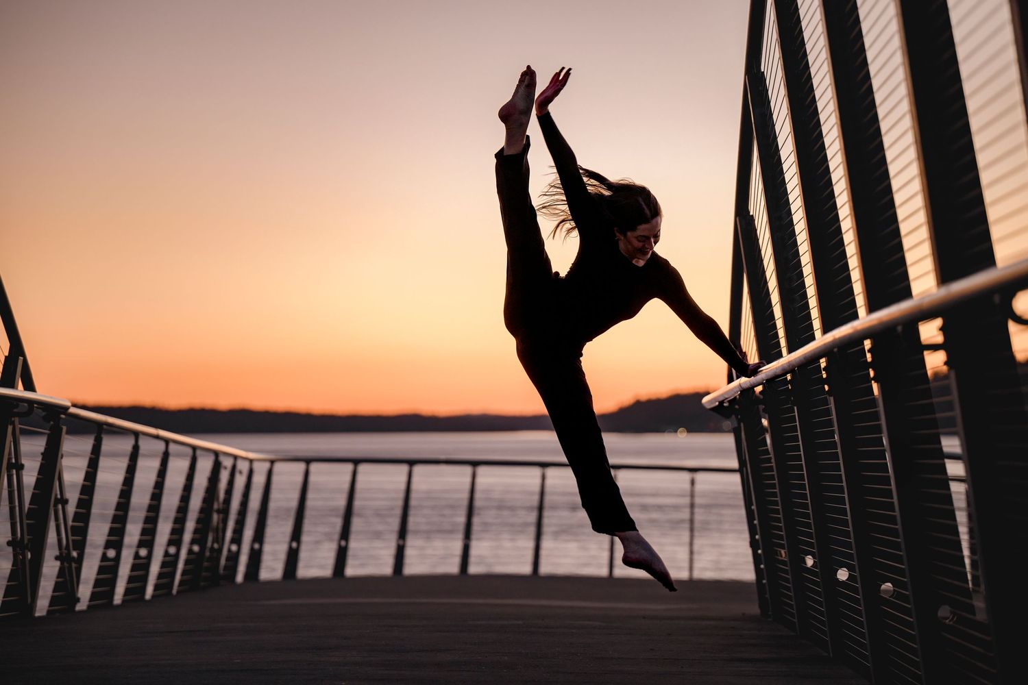 Silhouette of a female dancer performing a high kick split jump on a bridge at sunset with water in the background.