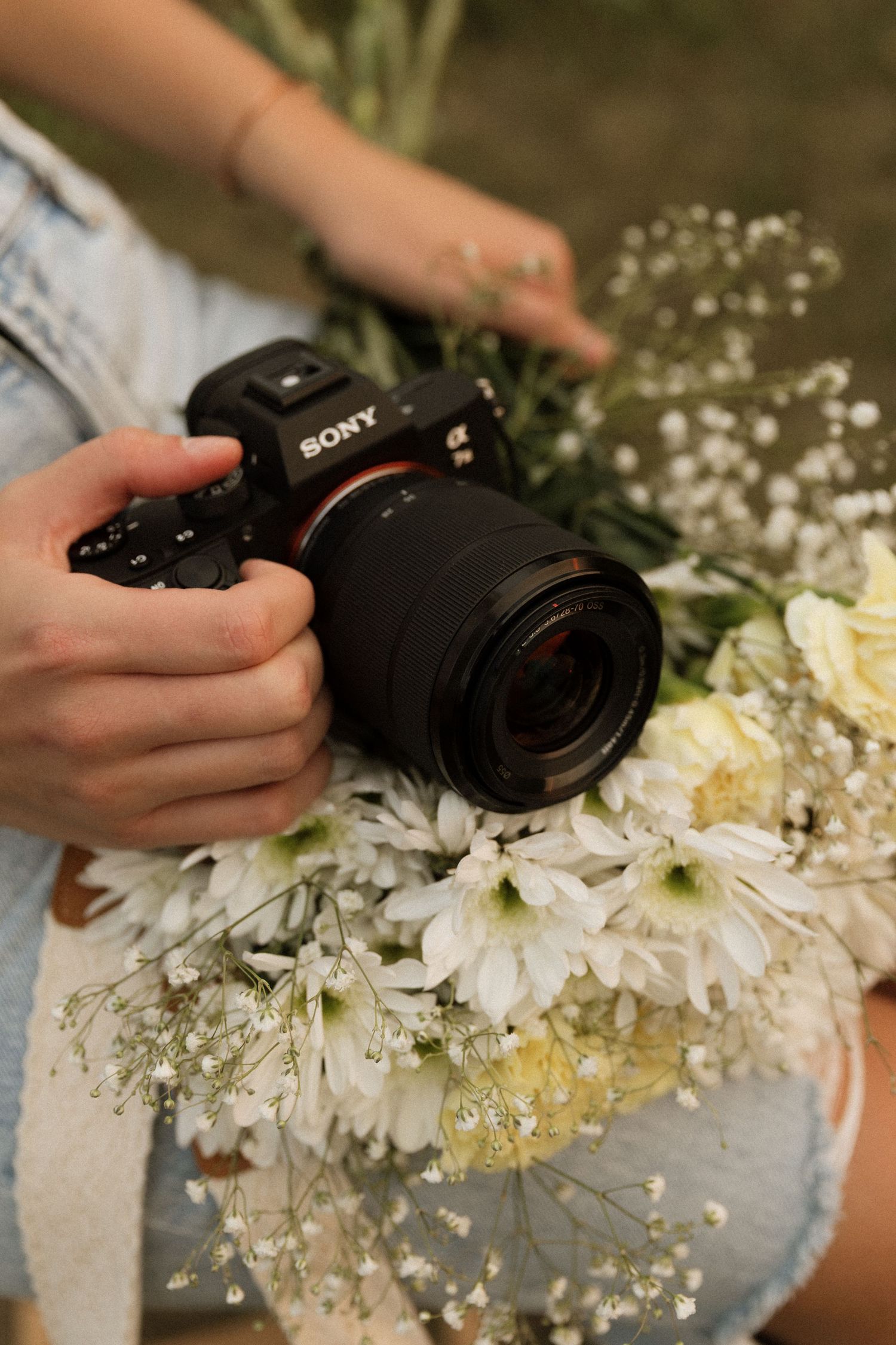 Hands holding Sony alpha camera resting on white flower bouquet outdoors.