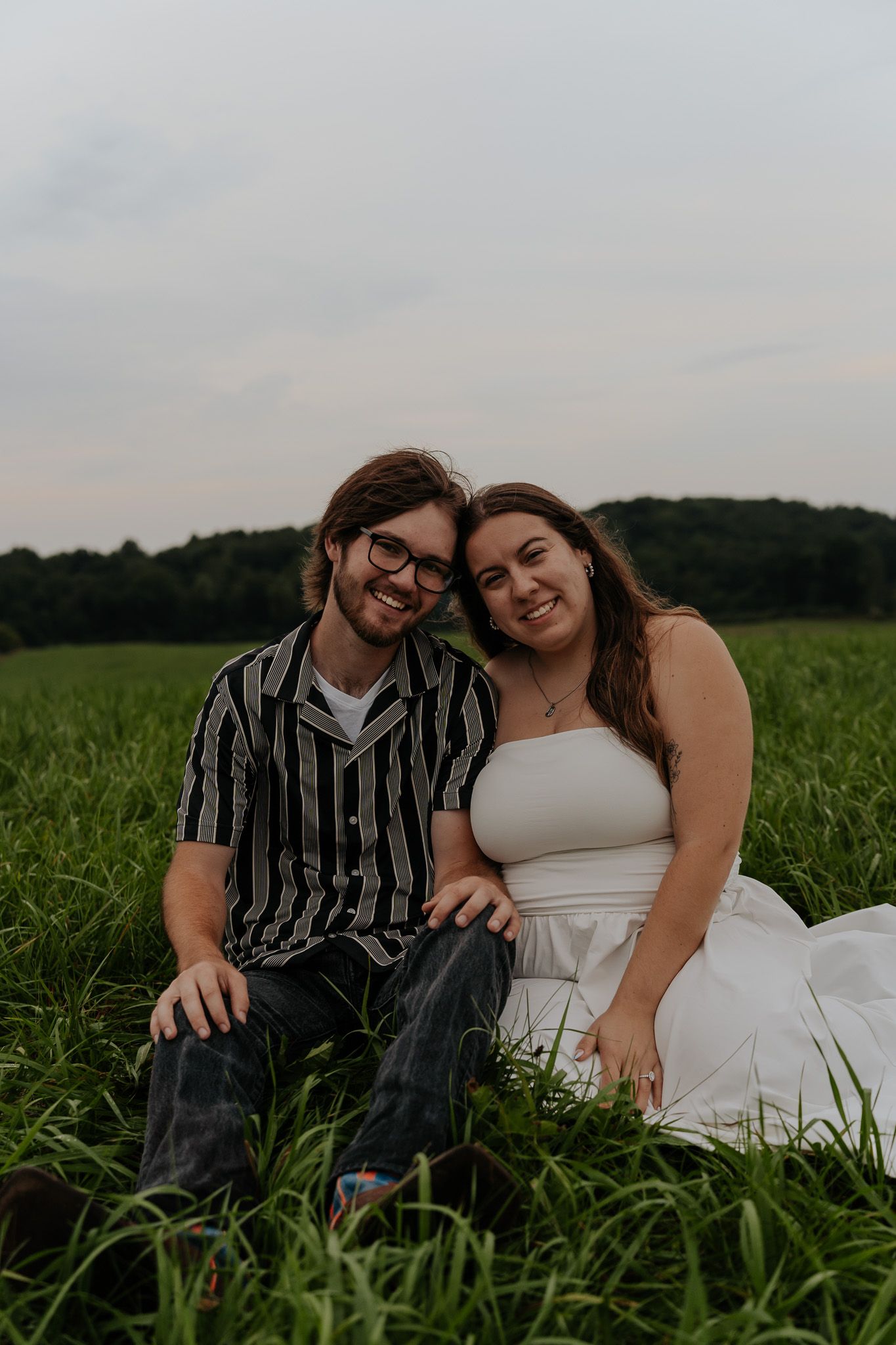Smiling engaged couple sitting together in tall green grass field at dusk during outdoor engagement photo session.