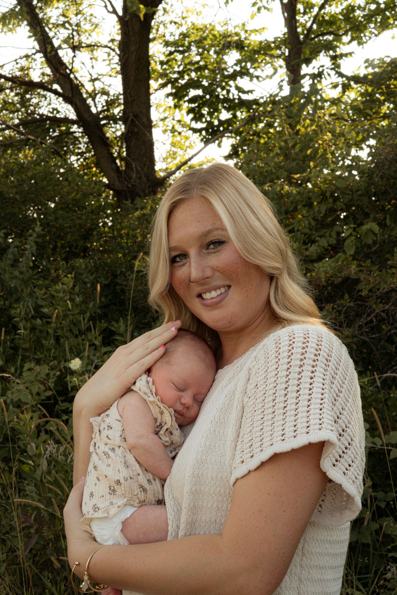 Smiling blonde mother holding sleeping newborn baby outdoors among green trees.