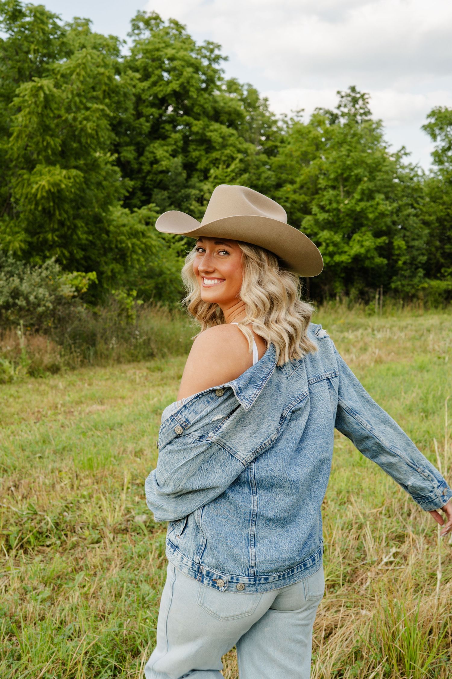 Cheerful blonde woman in tan cowboy hat and denim jacket twirling in a lush green meadow.