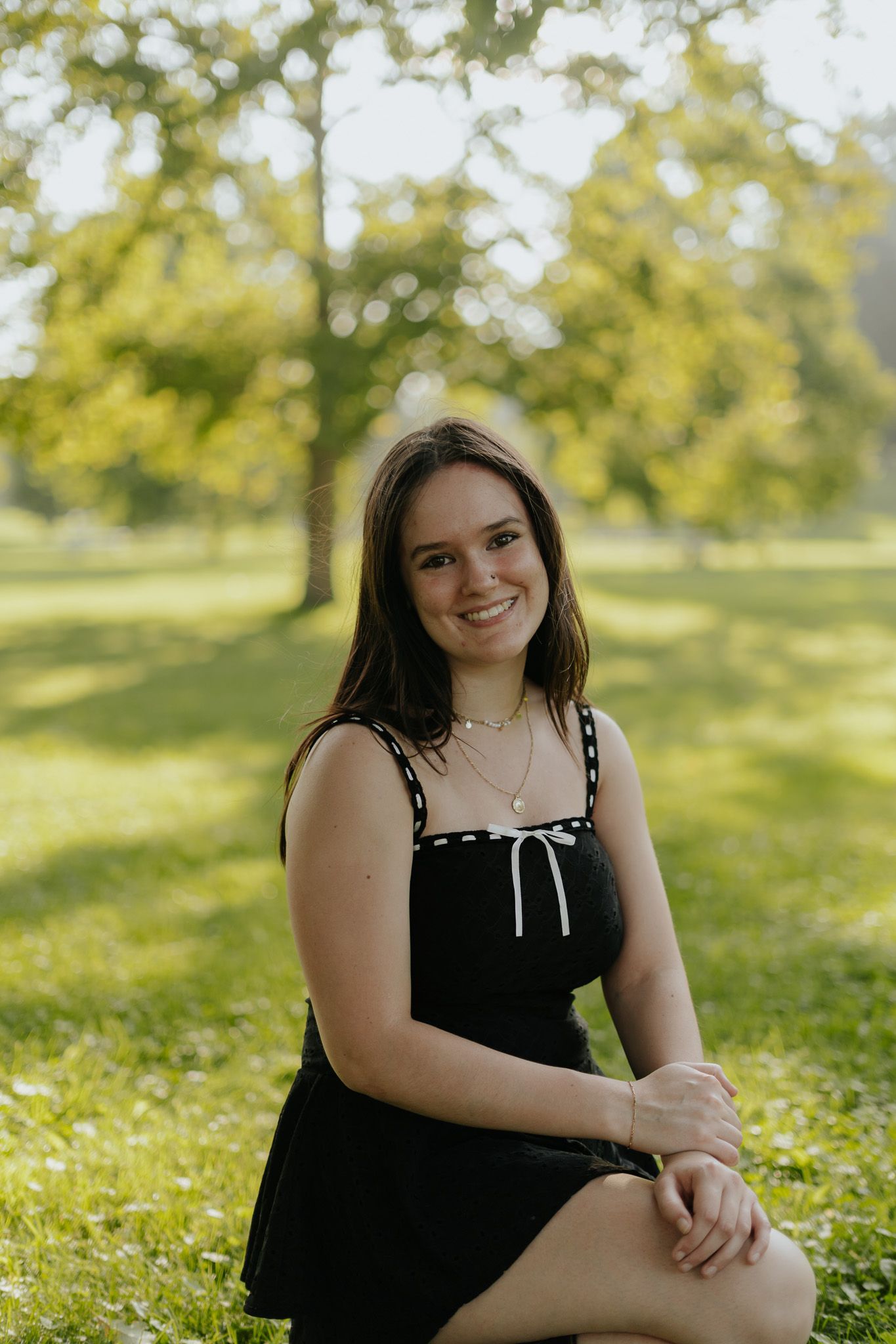 Smiling young woman in black sundress sitting on grass in sunny park.