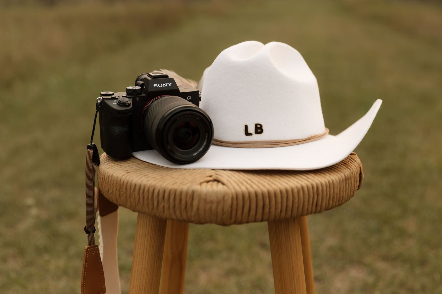 White cowboy hat with LB initials and Sony camera resting on wooden stool in grassy field.