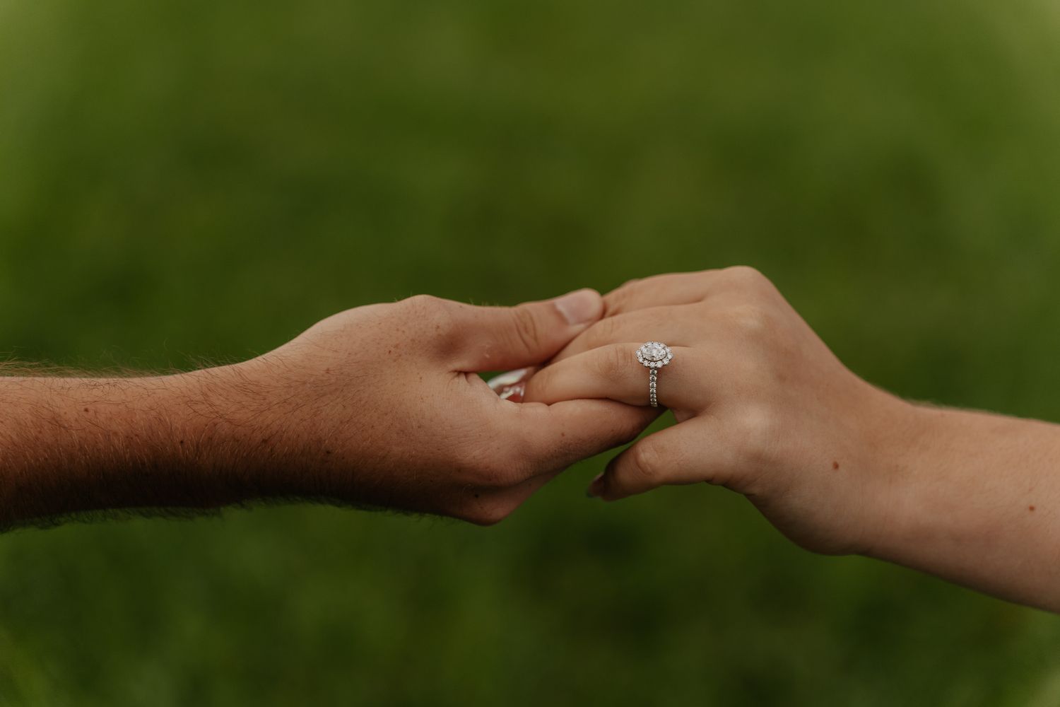 Close-up of couple holding hands in green field showcasing stunning halo diamond engagement ring on woman's finger.