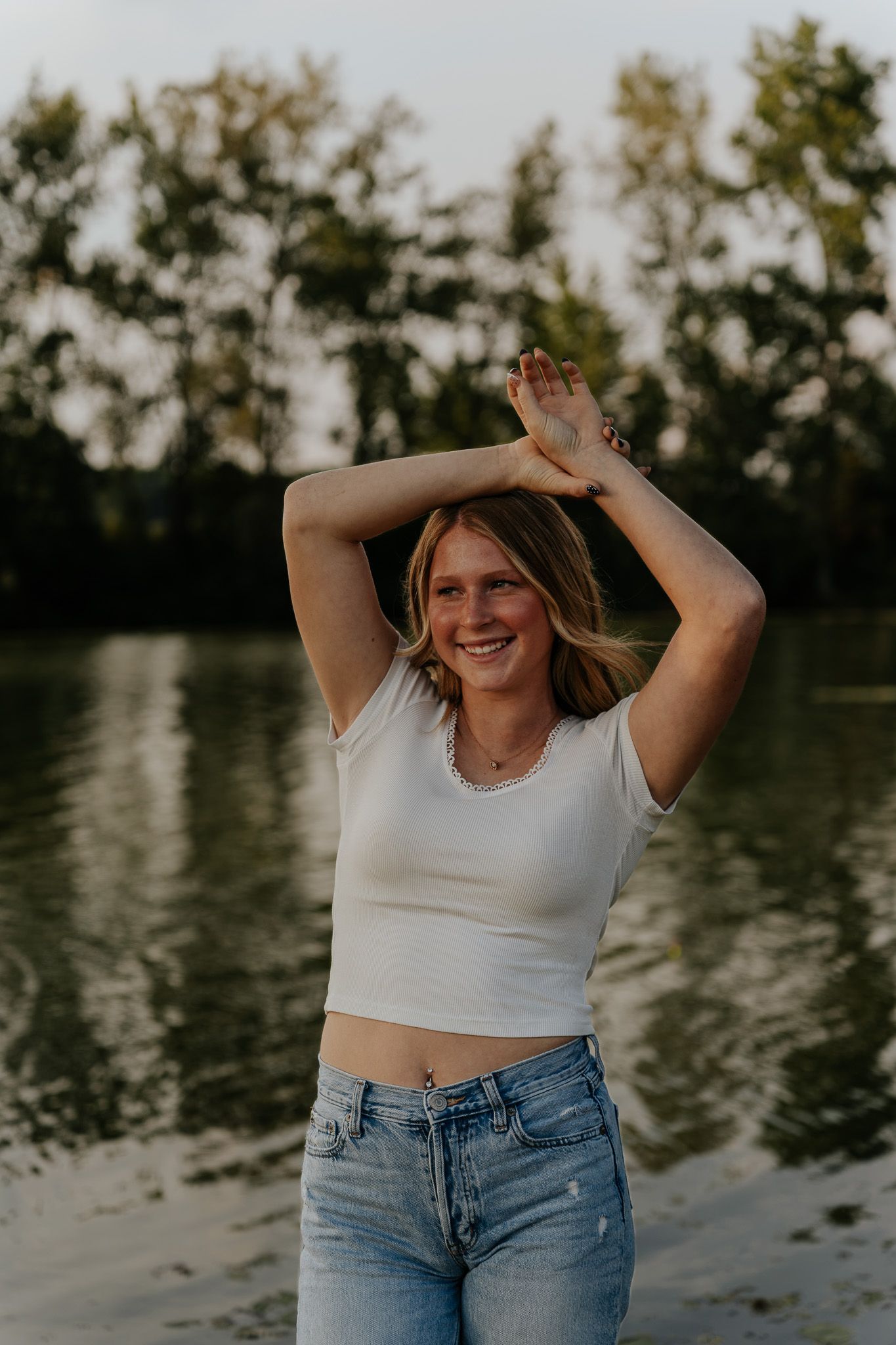 Smiling blonde woman in white crop top and jeans posing with arms raised near a peaceful lake.