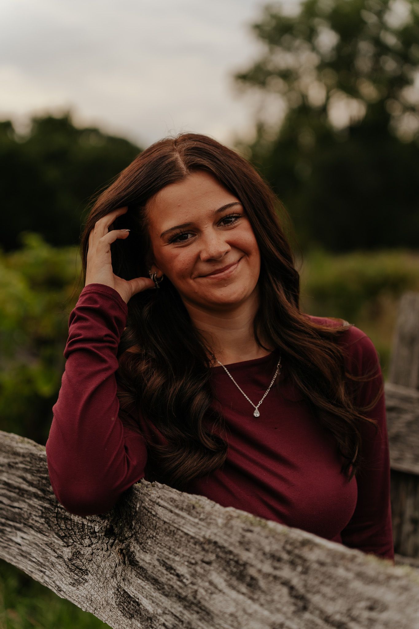 Young woman in burgundy top smiling while leaning on wooden fence.
