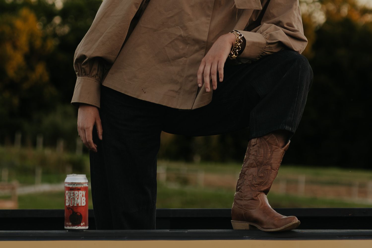Woman in tan jacket and cowboy boots with a can beside her on a ledge.