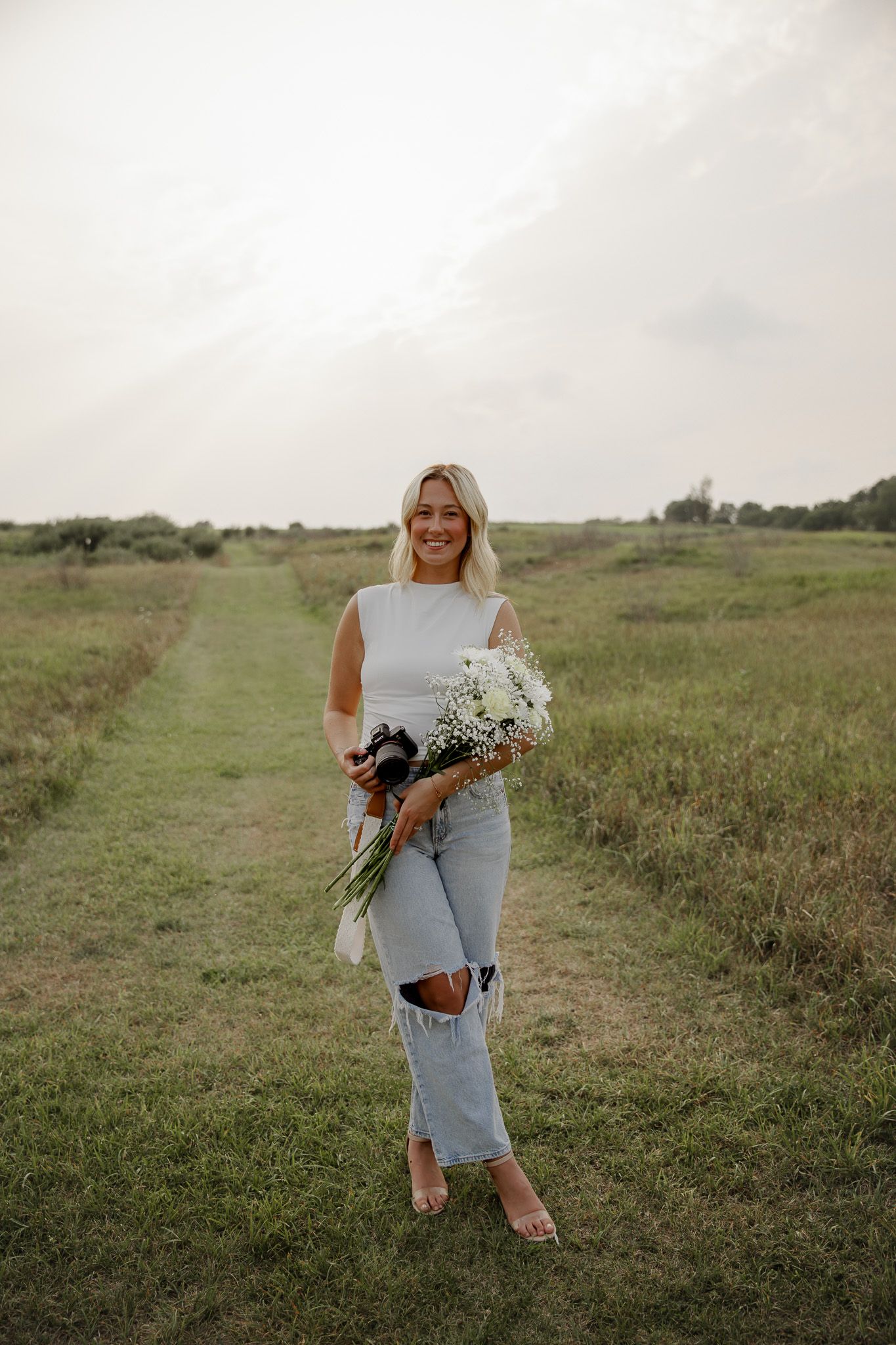 Smiling blonde woman holding white flowers and camera in an open green field.