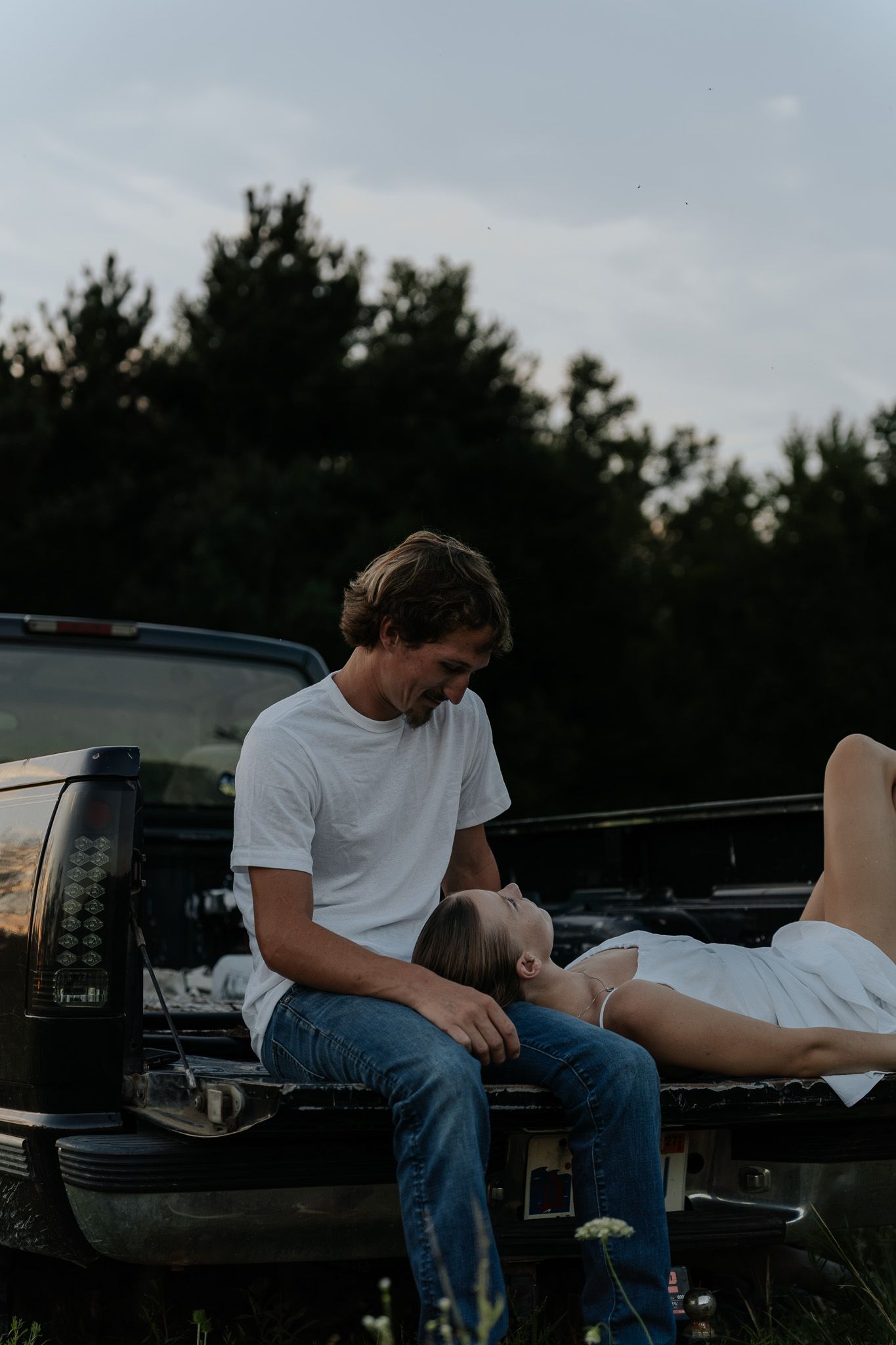 A man gazes lovingly at a woman lying across his lap on a truck tailgate at dusk.