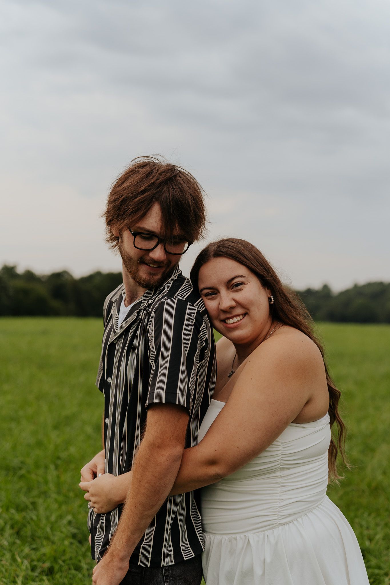 Joyful engaged couple embracing in green field, woman in white dress laughing while holding man in striped shirt.