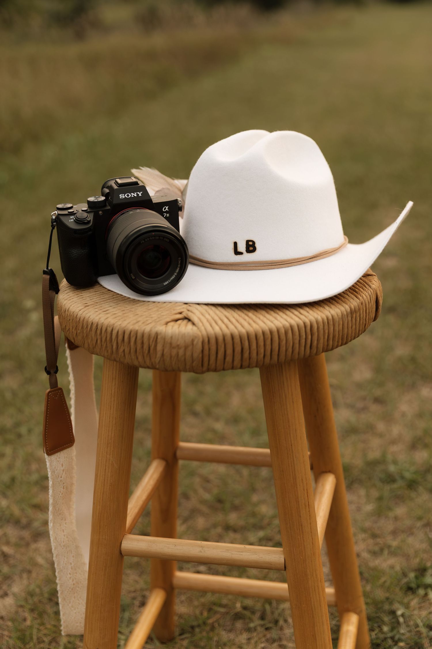 Sony camera and white cowboy hat with LB initials on wooden stool in field.