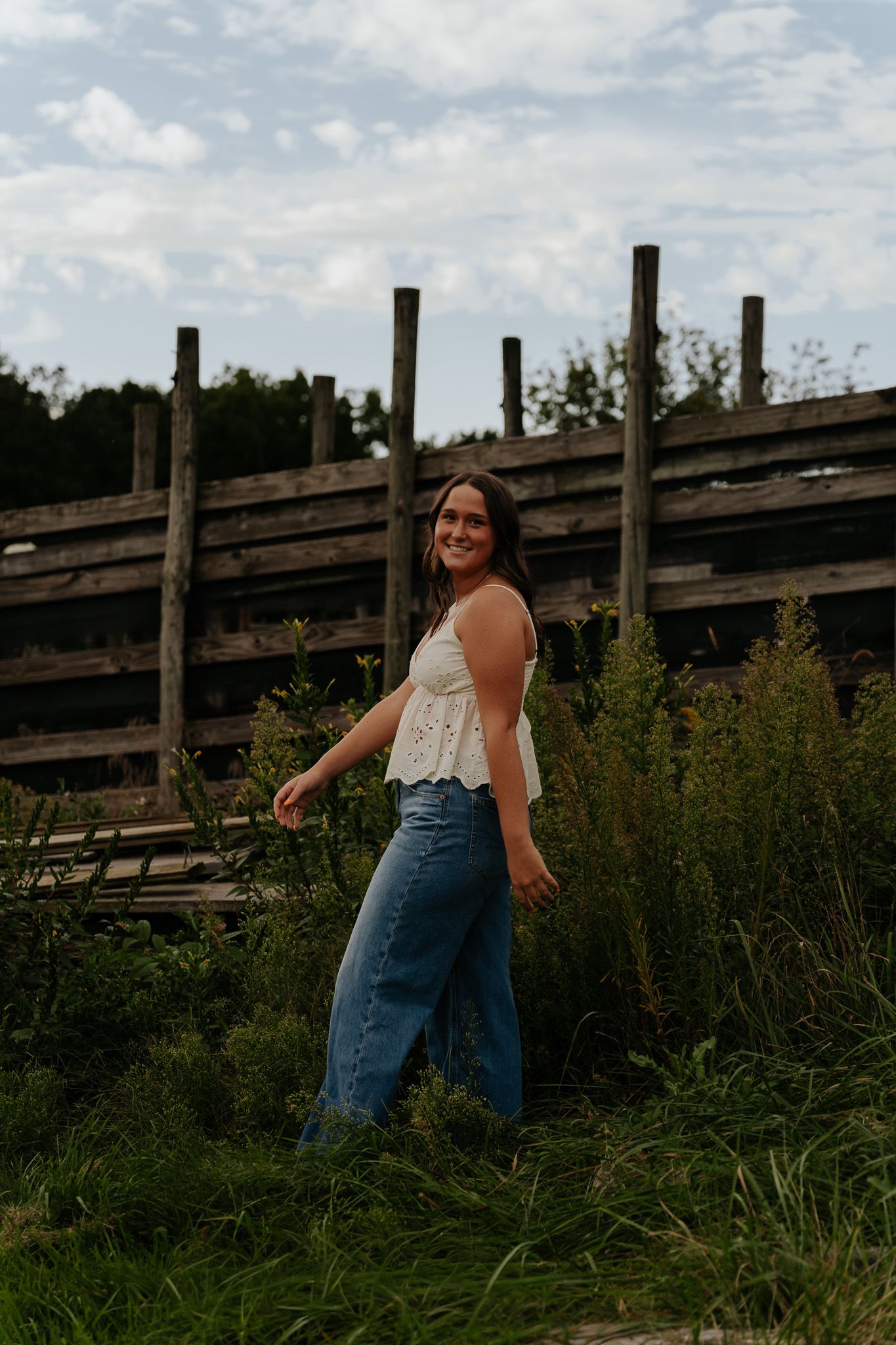 Smiling woman in white top and jeans walking through tall grass near wooden fence.