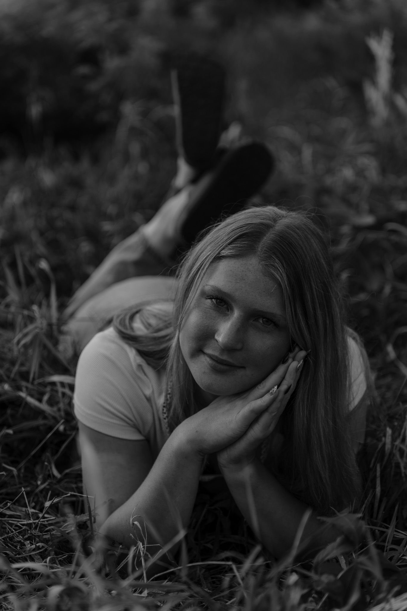 Black and white portrait of young blonde woman lying in grass looking thoughtfully at camera.