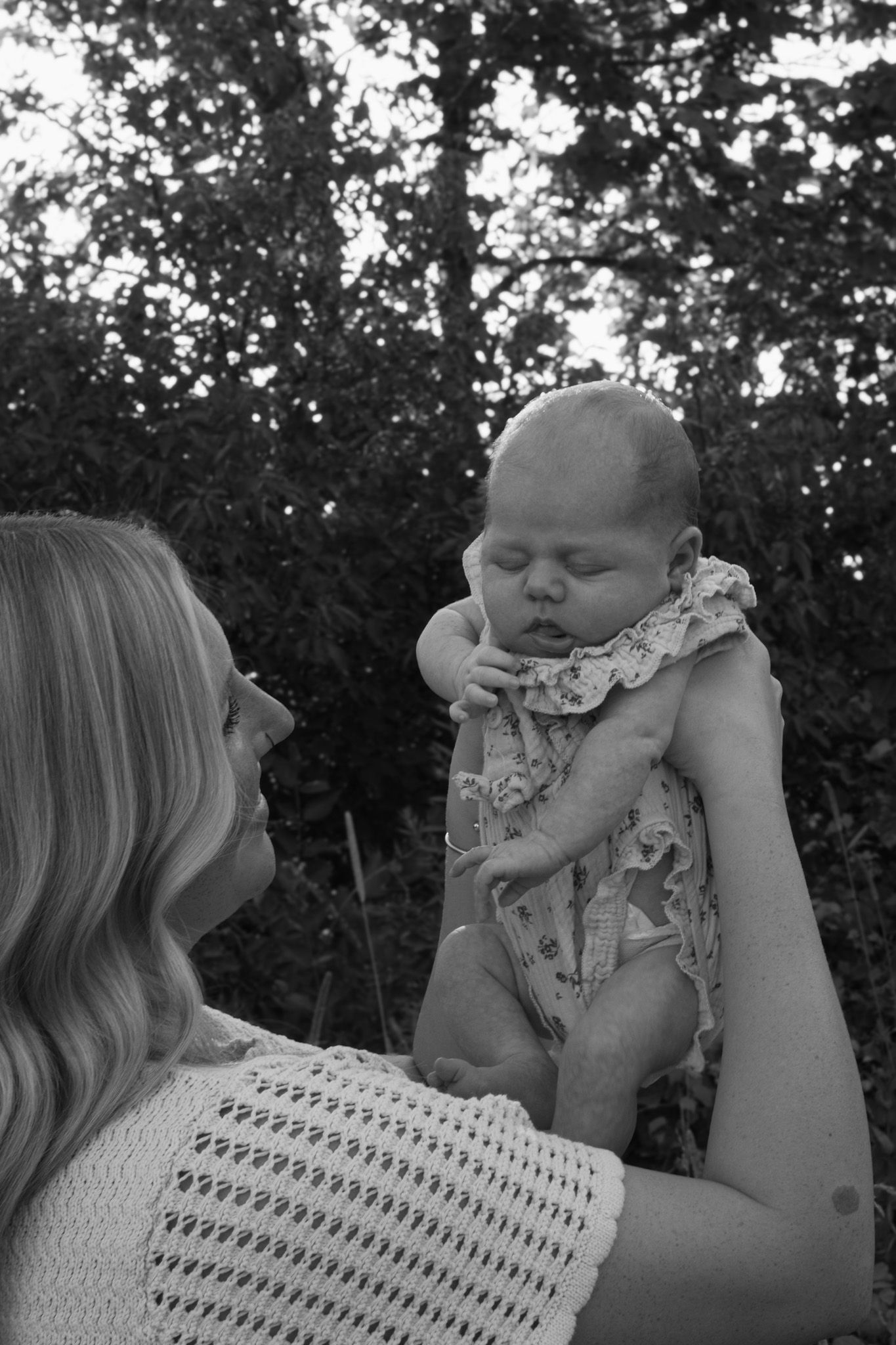 Black and white photo of a mother holding her newborn baby up in a lush green outdoor setting.