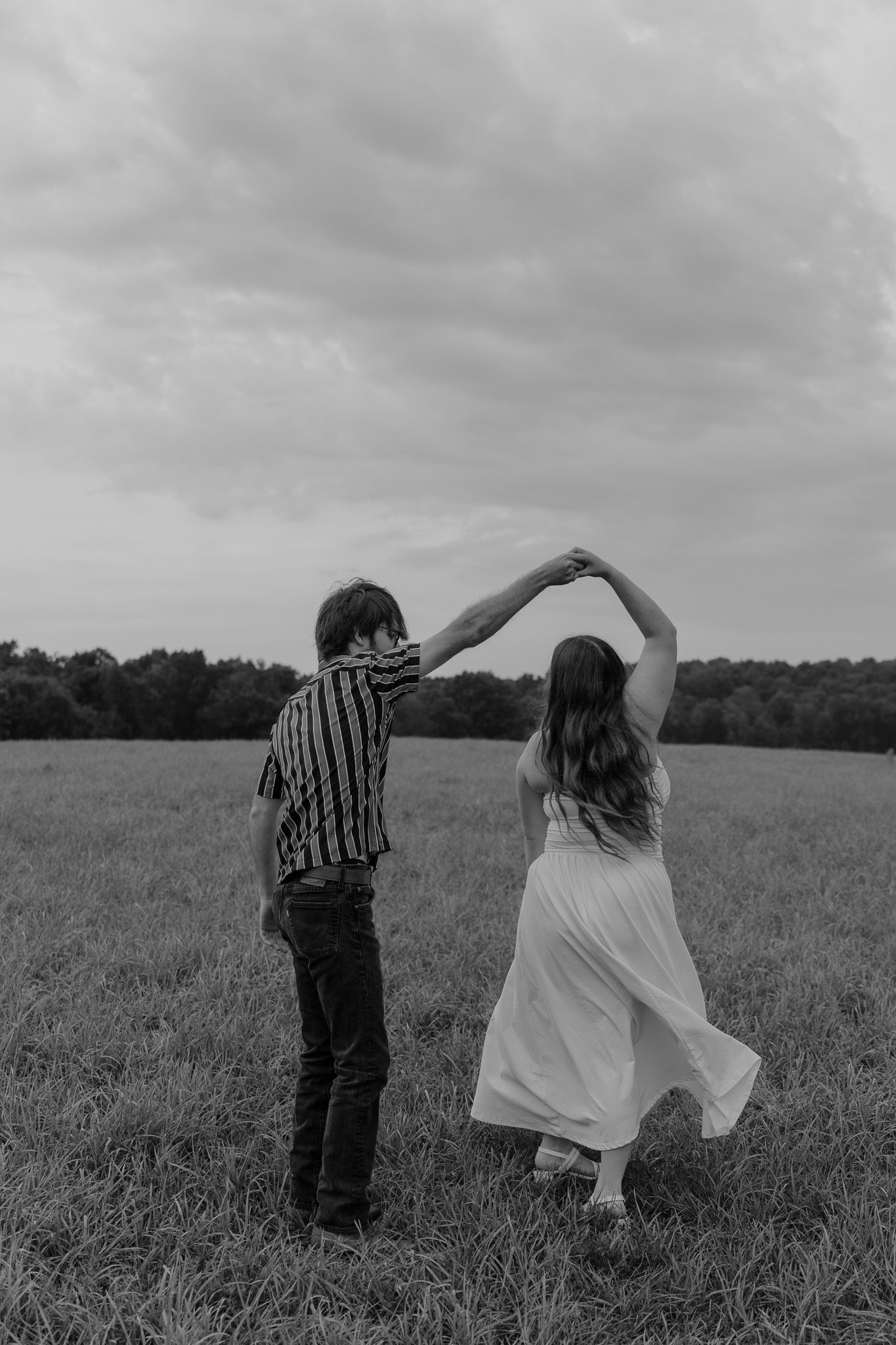 Black and white photo of engaged couple dancing in open field, man spinning woman in flowing white dress.