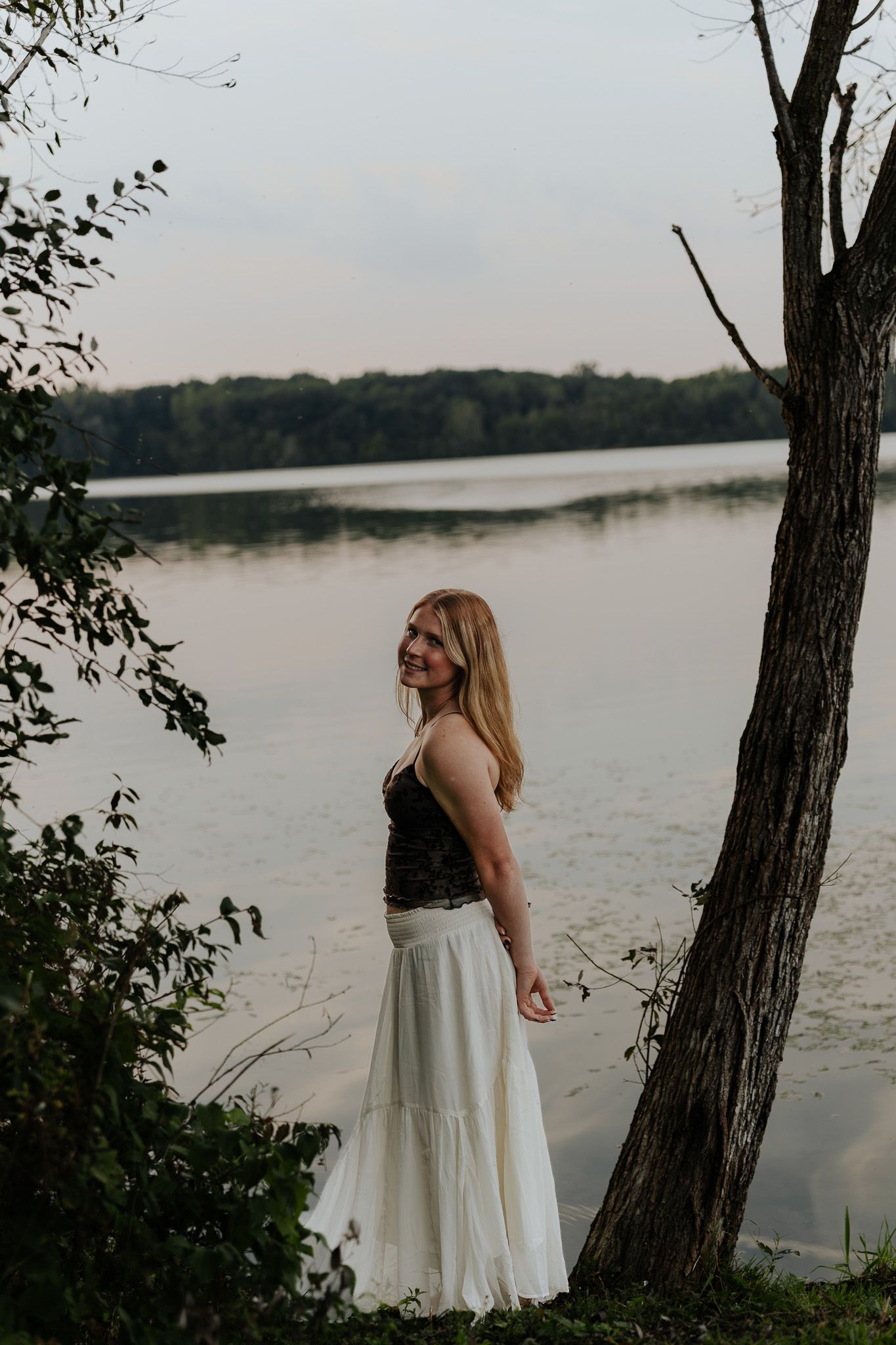 Smiling blonde woman in black top and white maxi skirt standing by tree near calm lake at dusk.