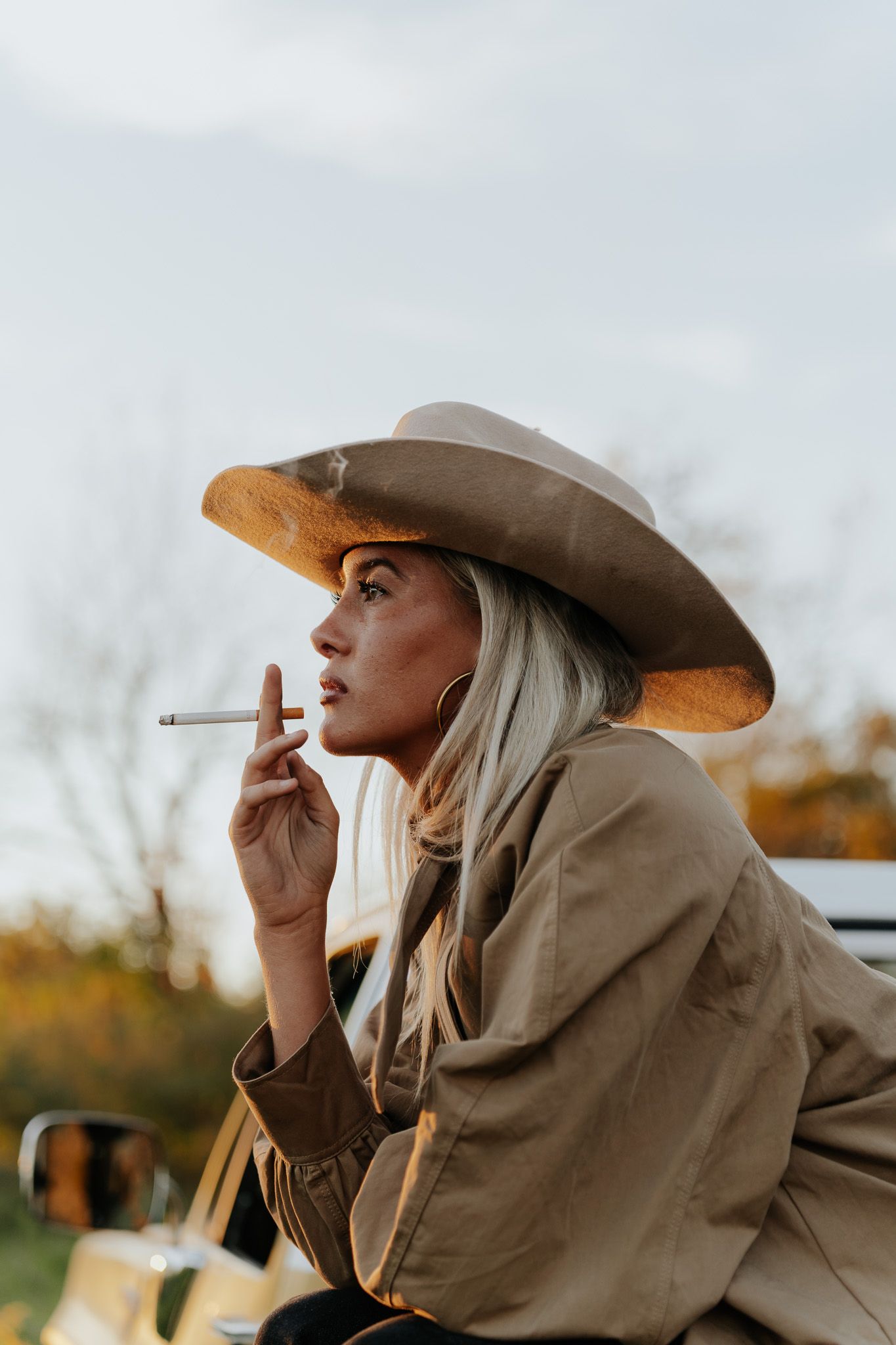 Blonde woman in cowboy hat and tan jacket smoking a cigarette outdoors at dusk.