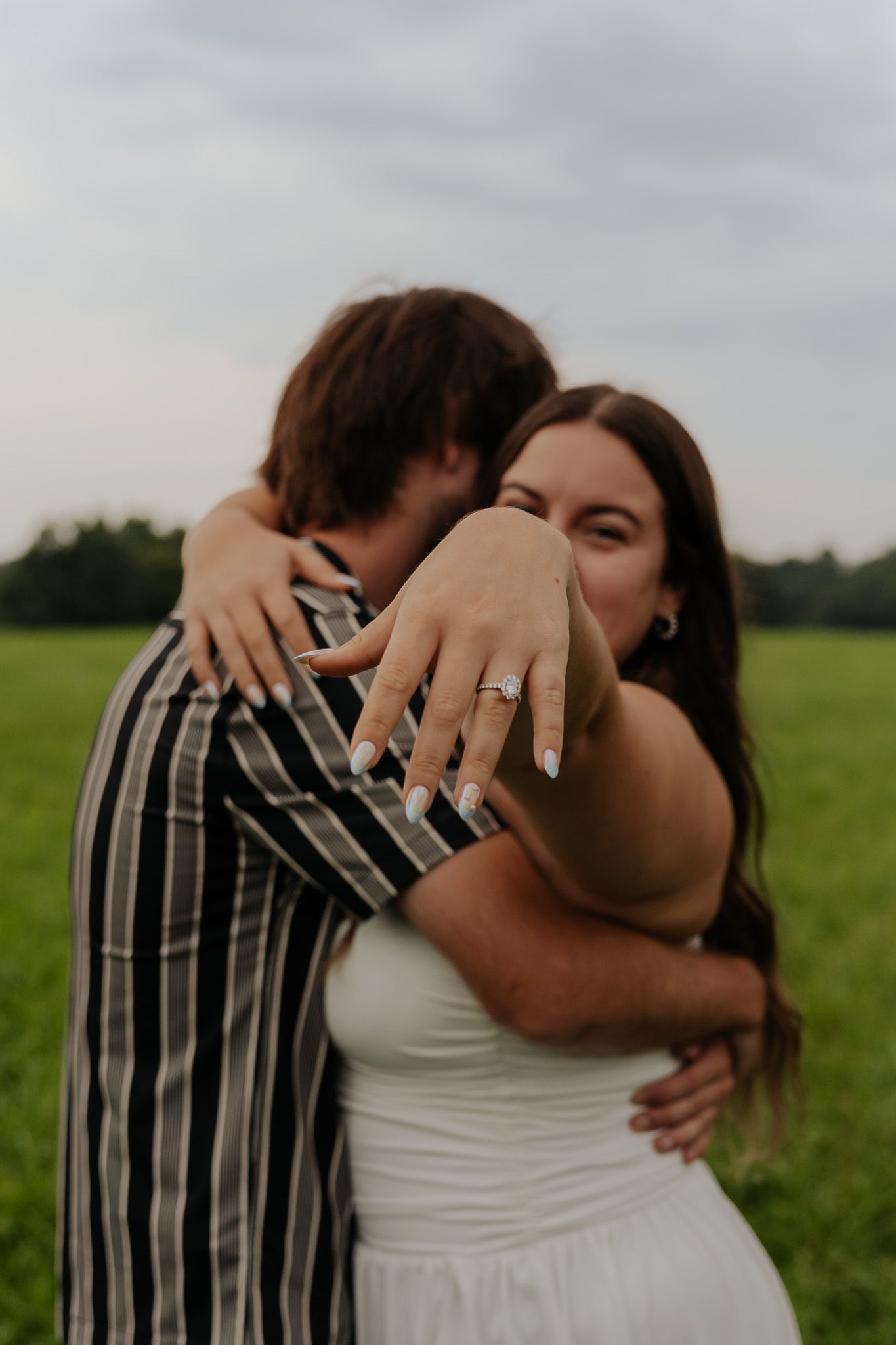 Bride-to-be hugging fiance in green field while showing off her sparkling halo diamond engagement ring to camera.