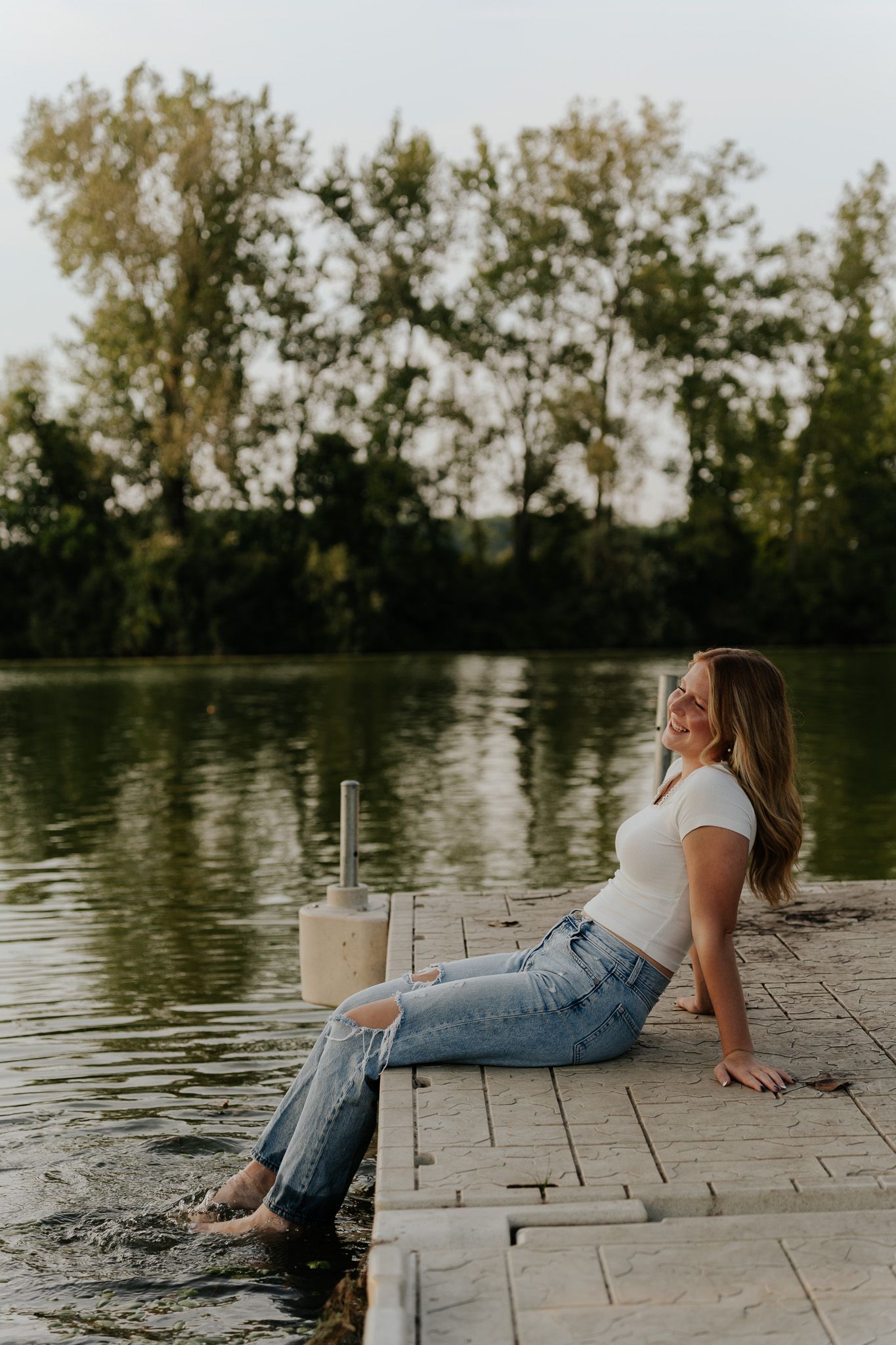 Blonde woman in white top and ripped jeans sitting on dock with feet in lake at sunset.
