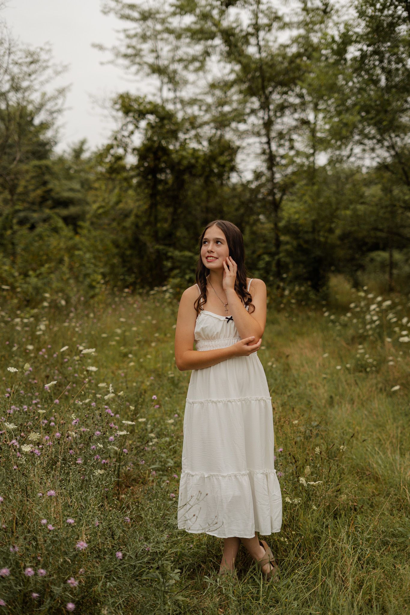 Young woman in white tiered sundress standing in wildflower meadow looking upward thoughtfully.