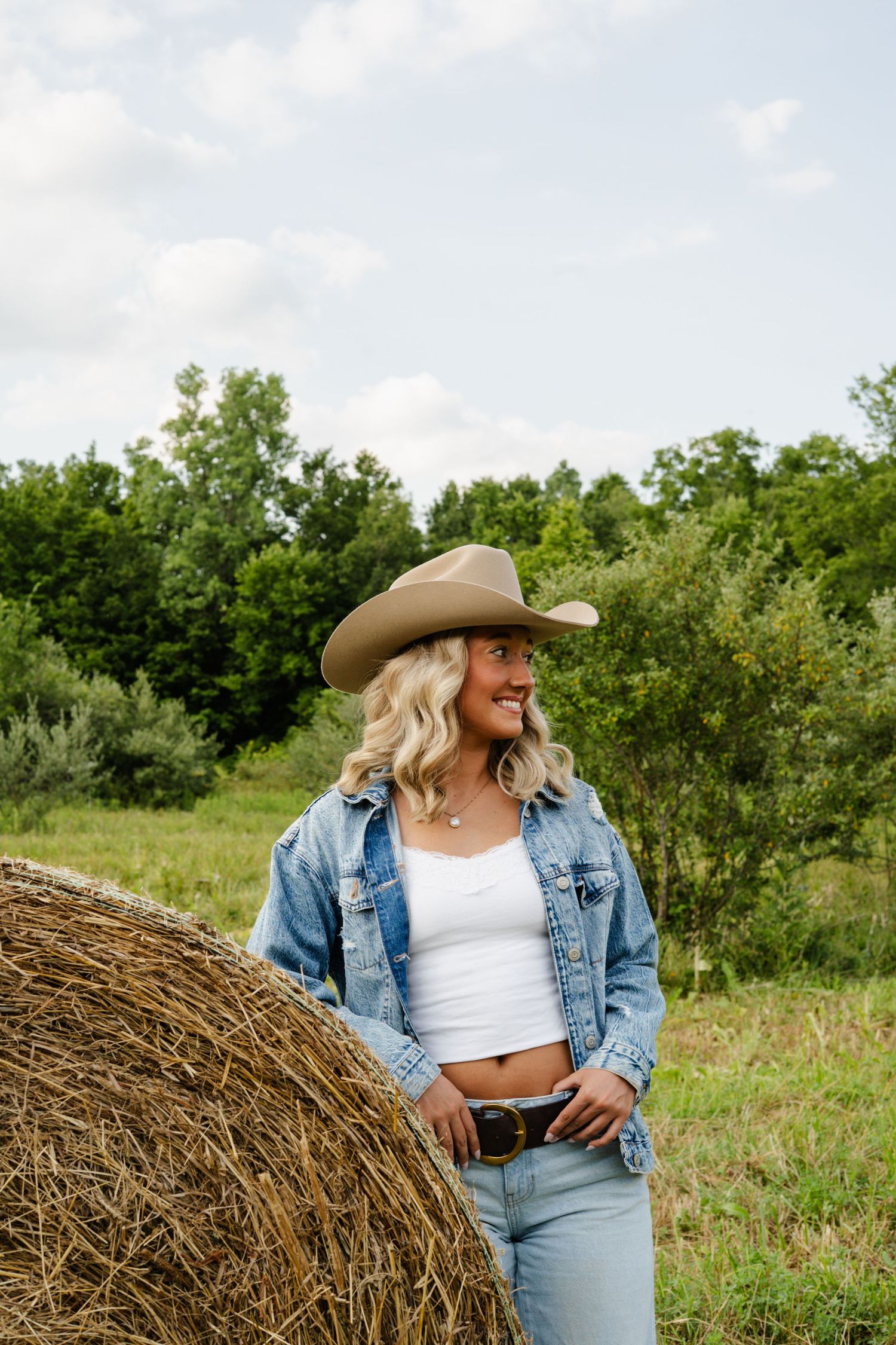 Smiling blonde woman in cowboy hat and denim jacket leaning against hay bale in green field.