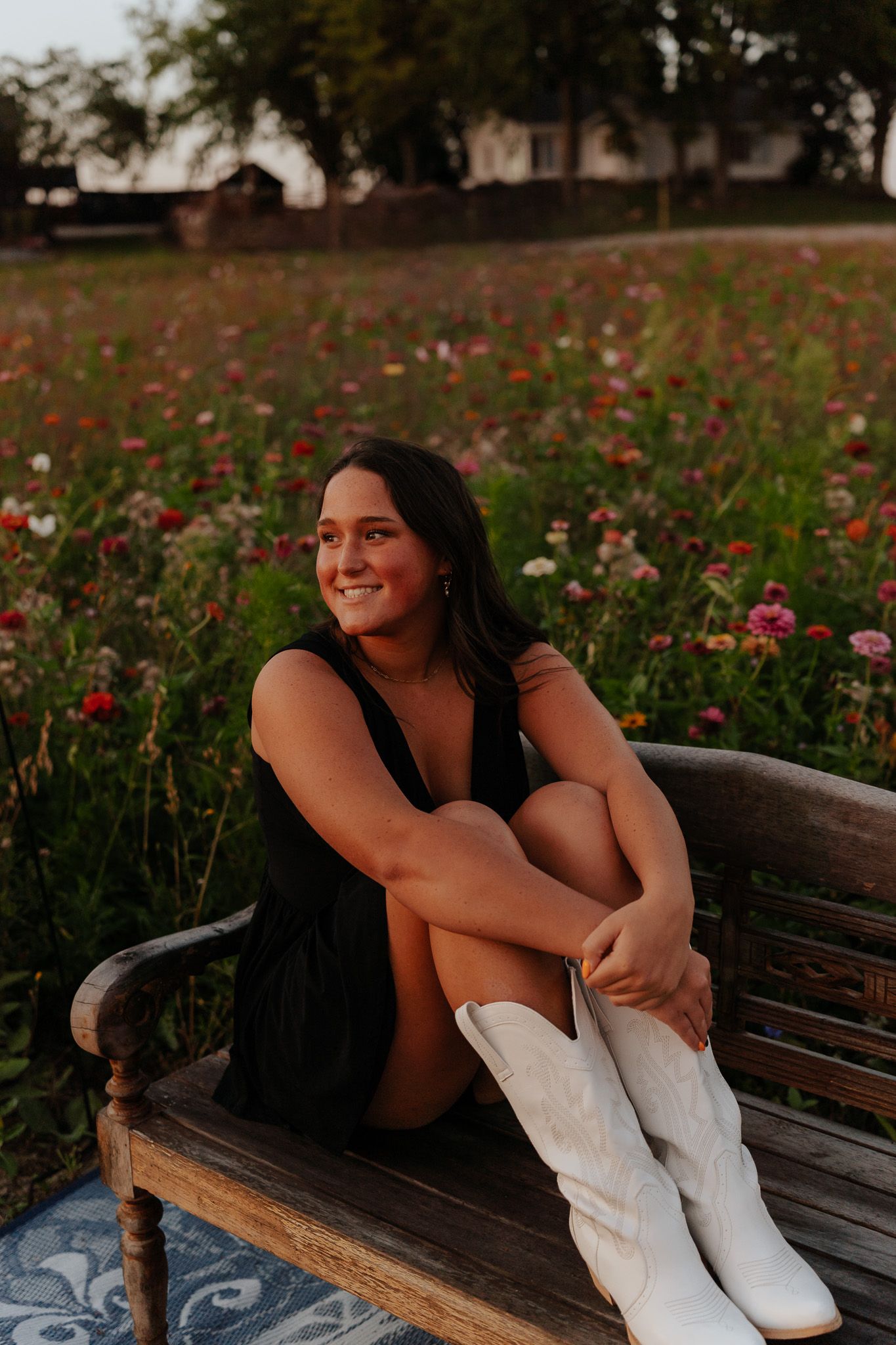 Smiling young woman in black dress and white cowboy boots sitting on bench in colorful wildflower field.
