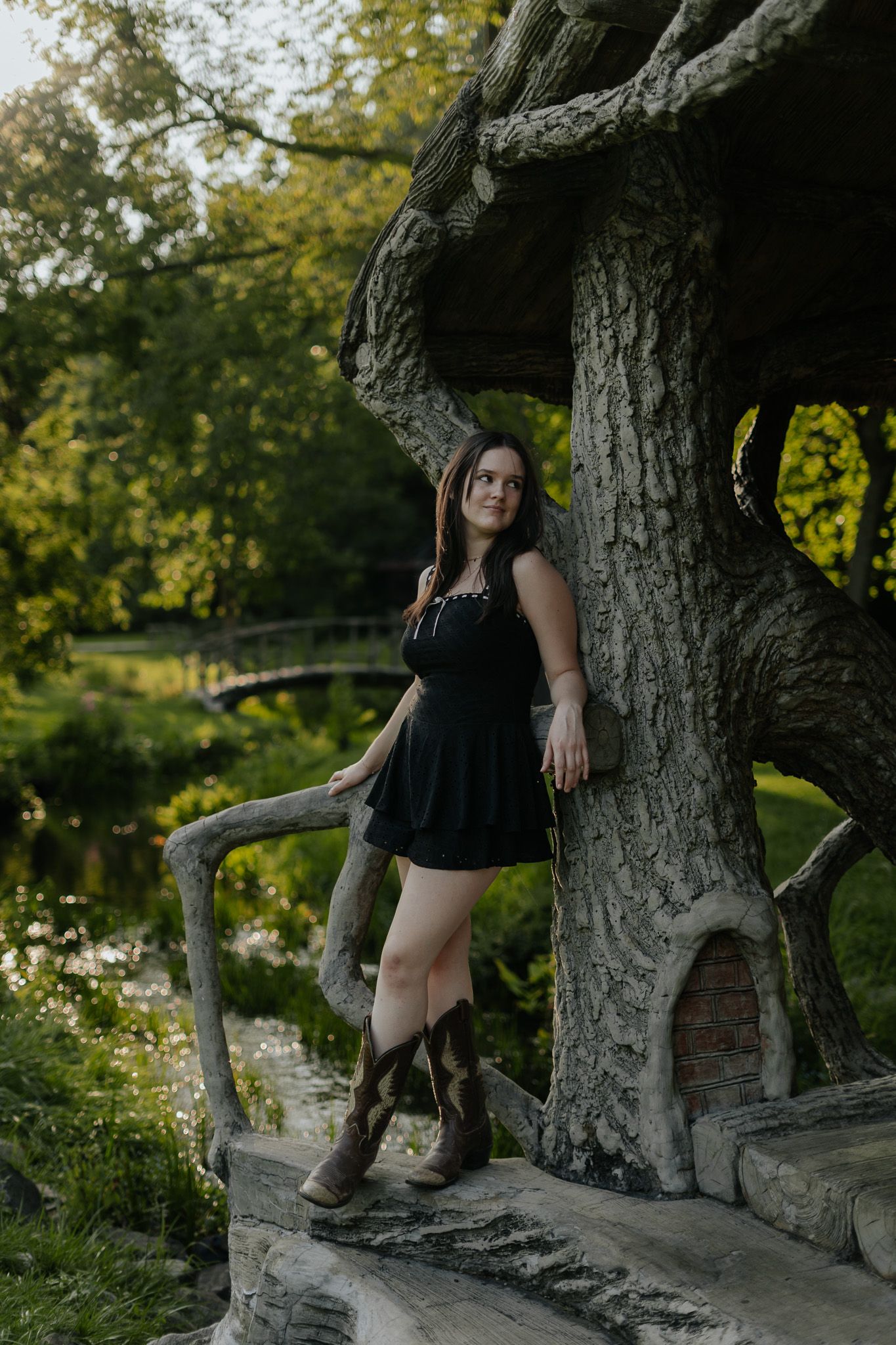 Woman in black dress and cowboy boots leaning against large tree in park.