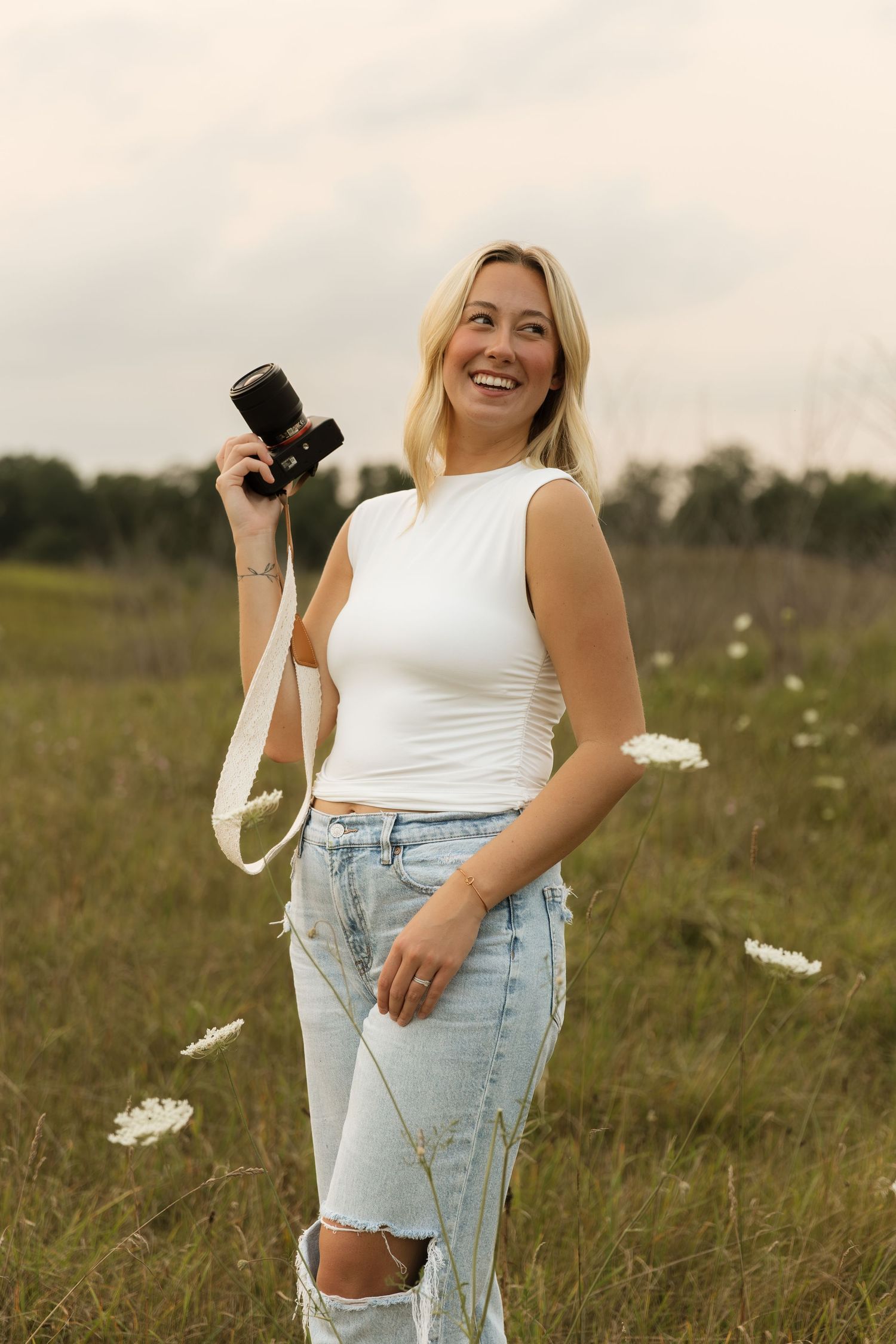 Laughing blonde woman holding Sony camera in white tank top and ripped jeans standing in open field.