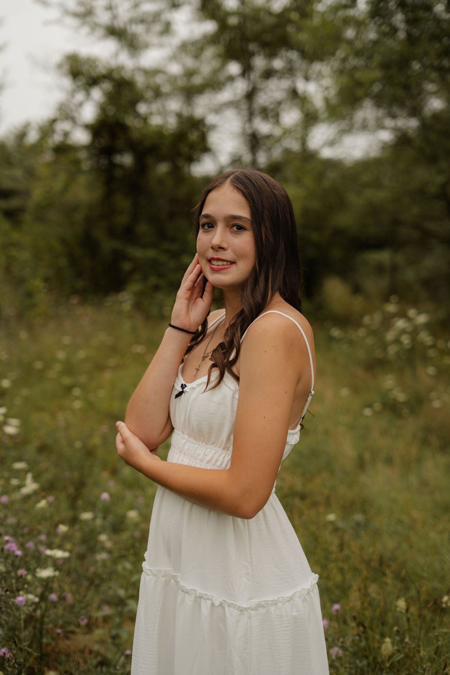 Smiling brunette teen in white dress posing in wildflower field with hand near her face.