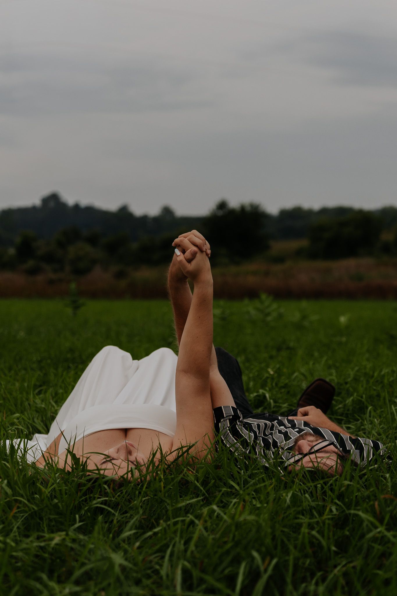 Engaged couple lying in lush green grass holding hands raised up toward cloudy sky in romantic outdoor session.