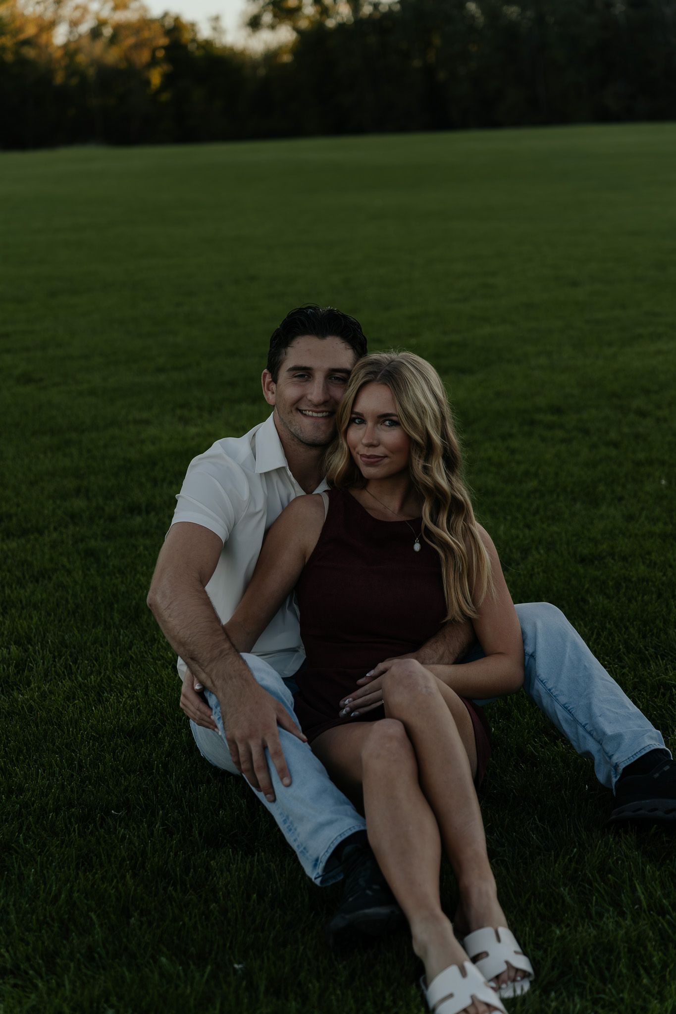 Couple sitting together on a lush green lawn, smiling warmly during their engagement session.