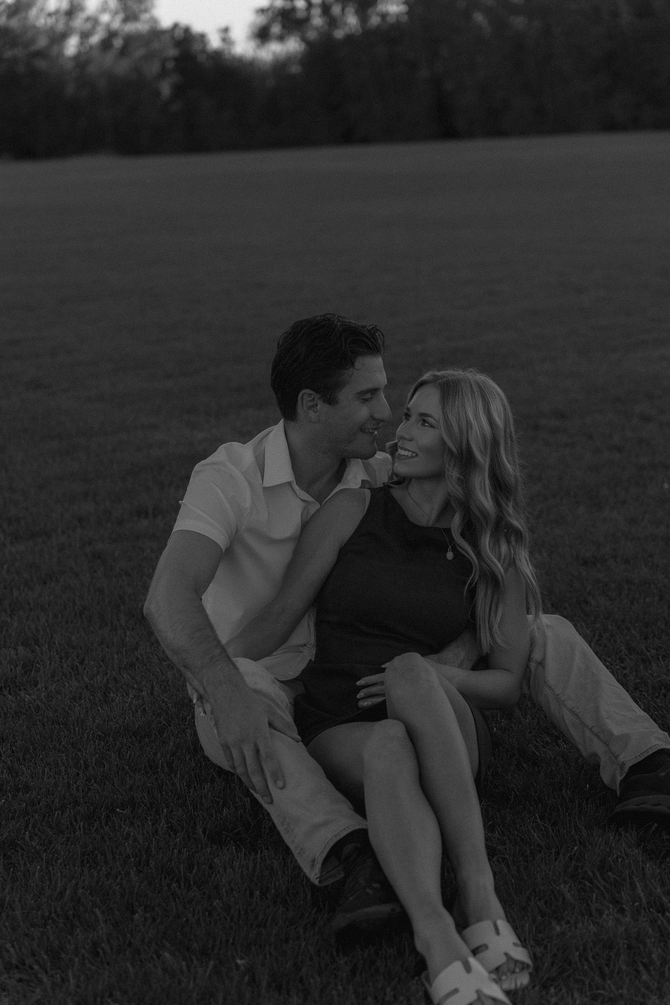Black and white photo of a smiling couple sitting together on a grassy field at sunset.